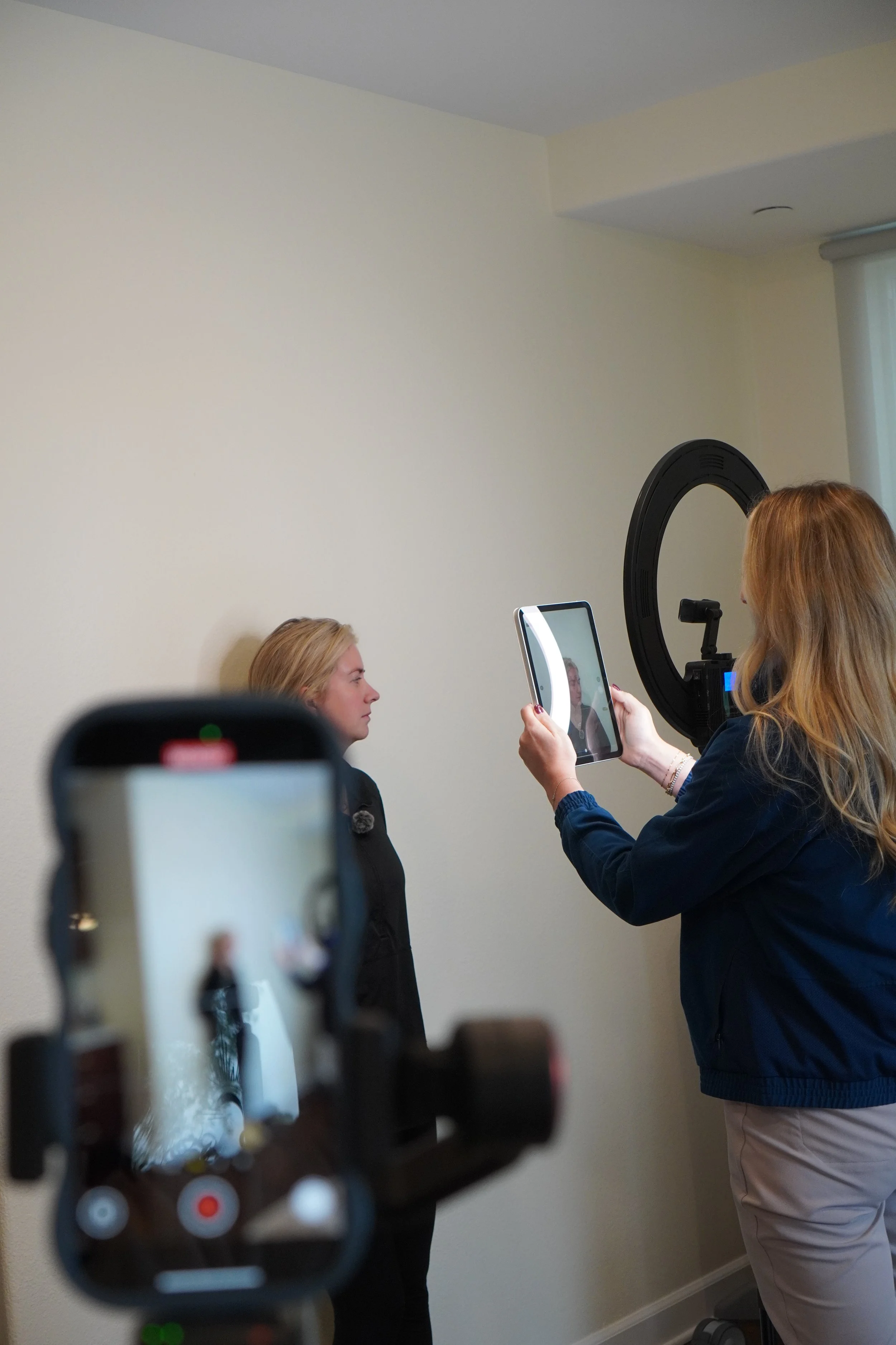 A woman with red hair is taking a portrait photo of a blonde woman against a plain wall using a tablet. A camera on a tripod is also capturing the scene.