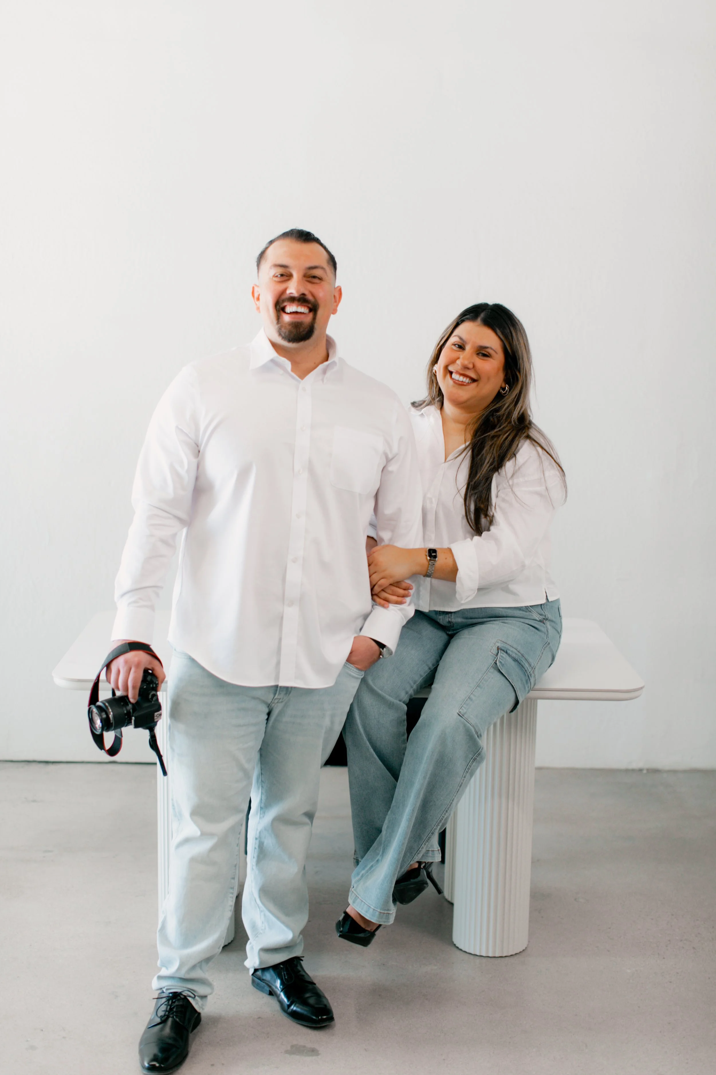 Two people smiling in casual white shirts, with one holding a camera, pose in front of a plain white background.