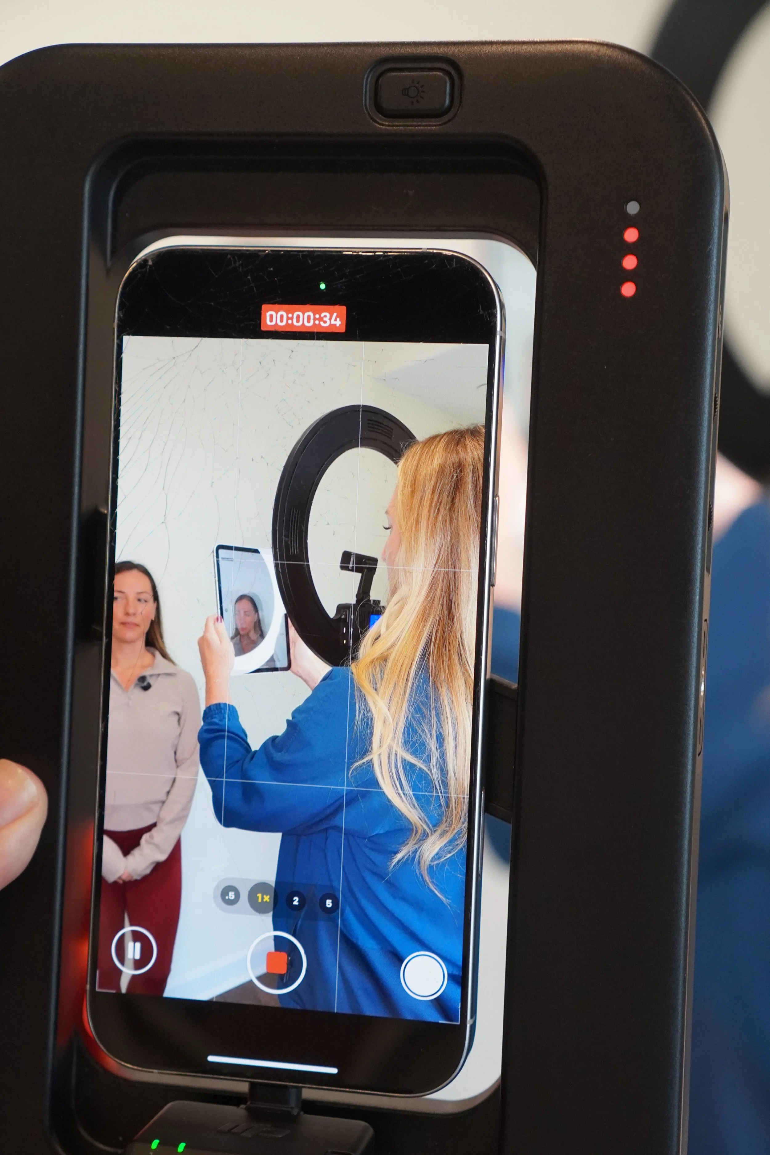 A woman is recording a video of another woman in front of a white wall with a mounted device, using a smartphone on a stabilizer or tripod, with a cracked screen visible in the video frame.
