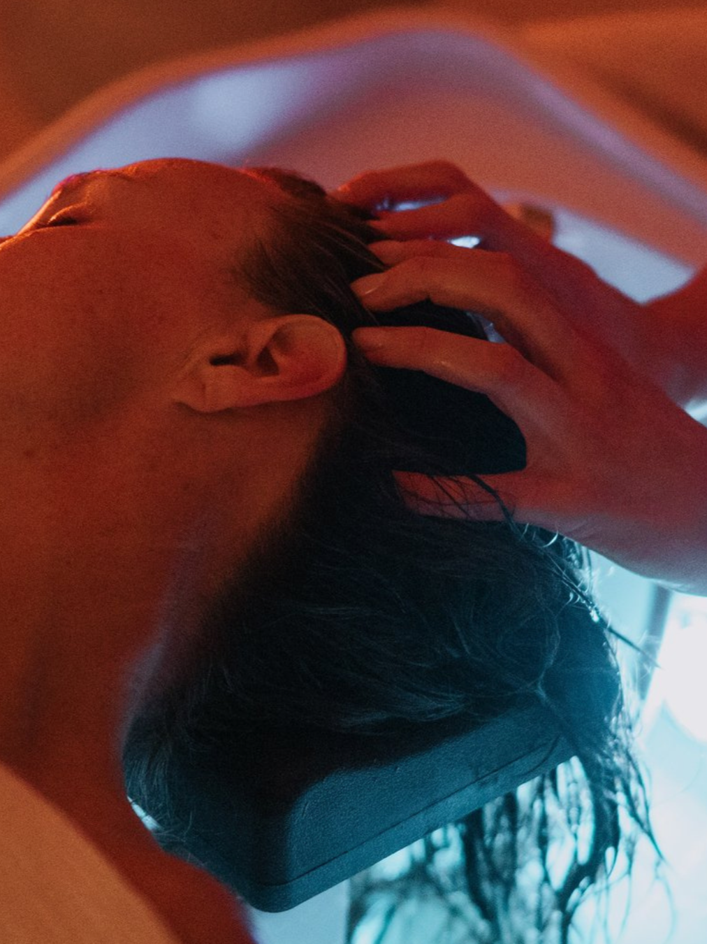 Une personne allongée sur une table, tête sur le côté, avec des cheveux longs et foncés, dans une ambiance tamisée et colorée.