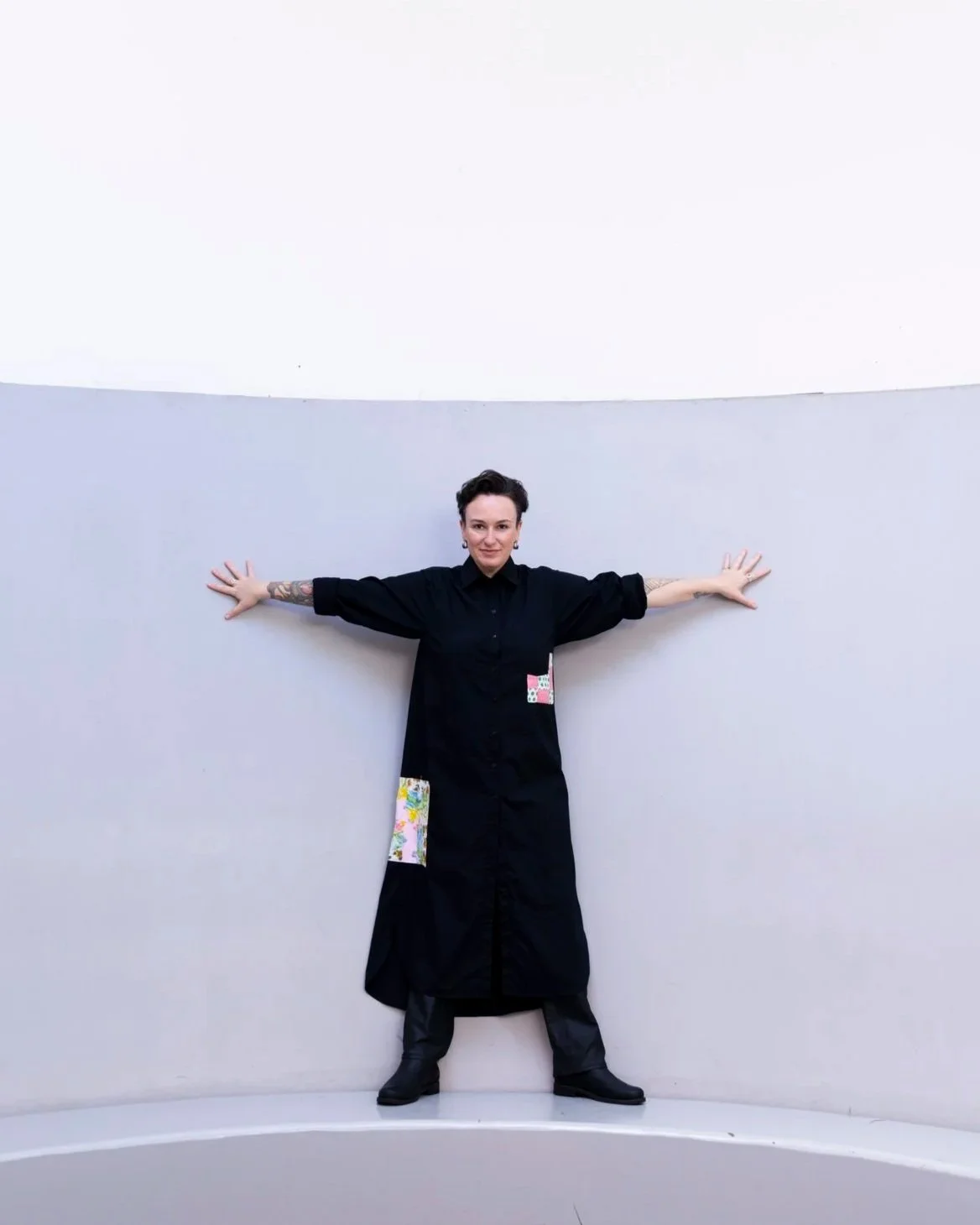 Person with short dark hair standing against a curved, high wall, arms outstretched, wearing a black long dress with colorful patches, and black boots.