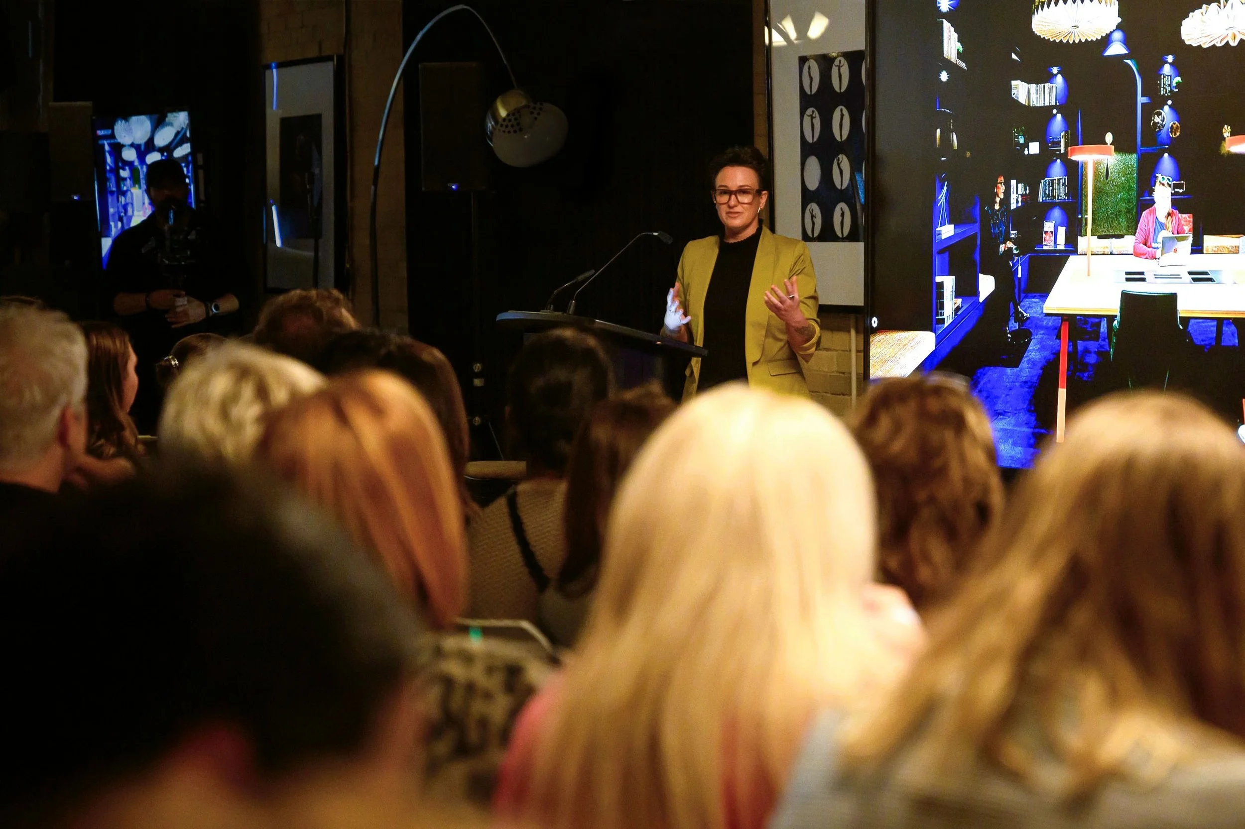 Person giving a presentation at a conference or seminar, audience members listening attentively, and a large screen displaying a room interior behind them.