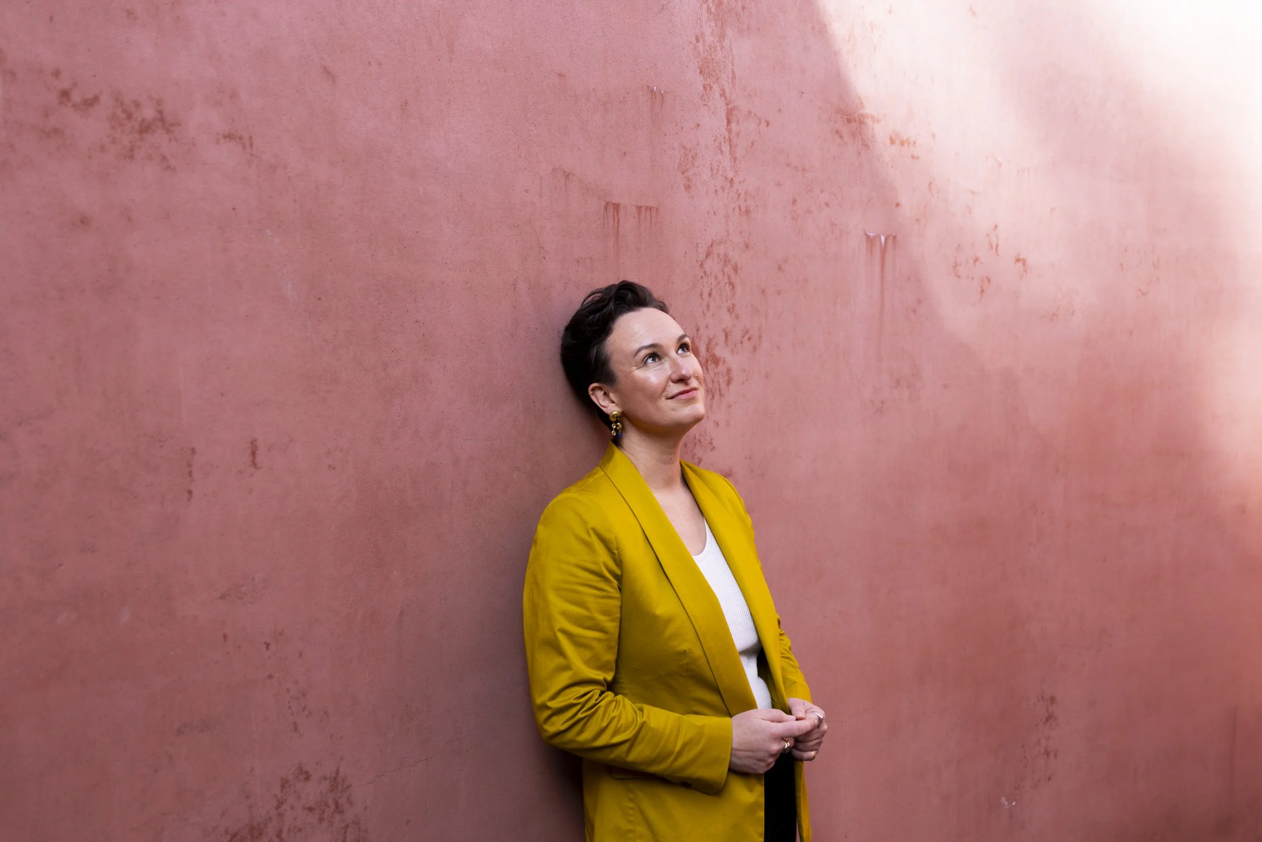 A person with short dark hair, wearing a yellow blazer and earrings, standing against a pink textured wall, looking thoughtfully upward.