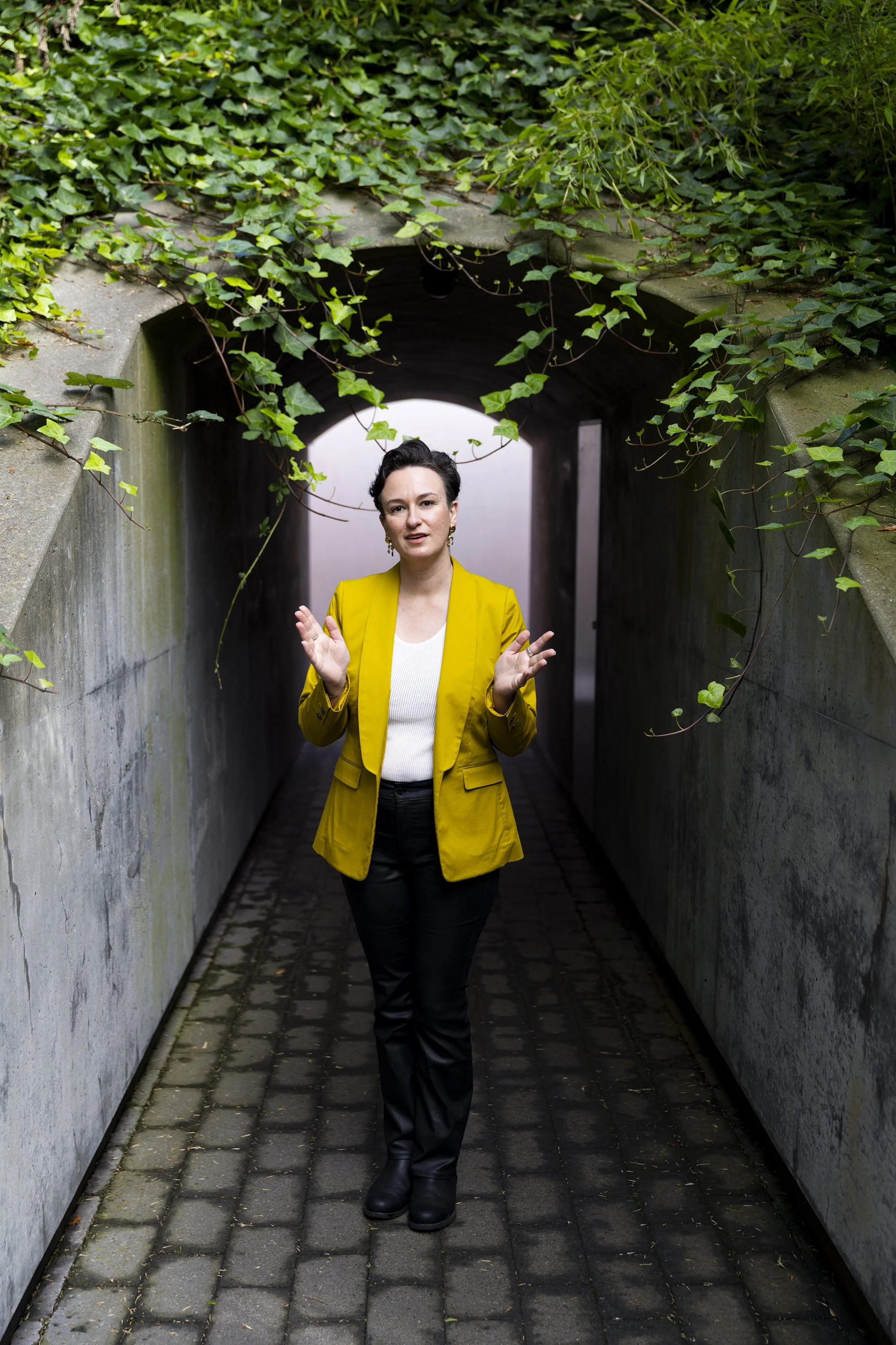Woman in a yellow blazer and black pants walking through a green, vine-covered tunnel.