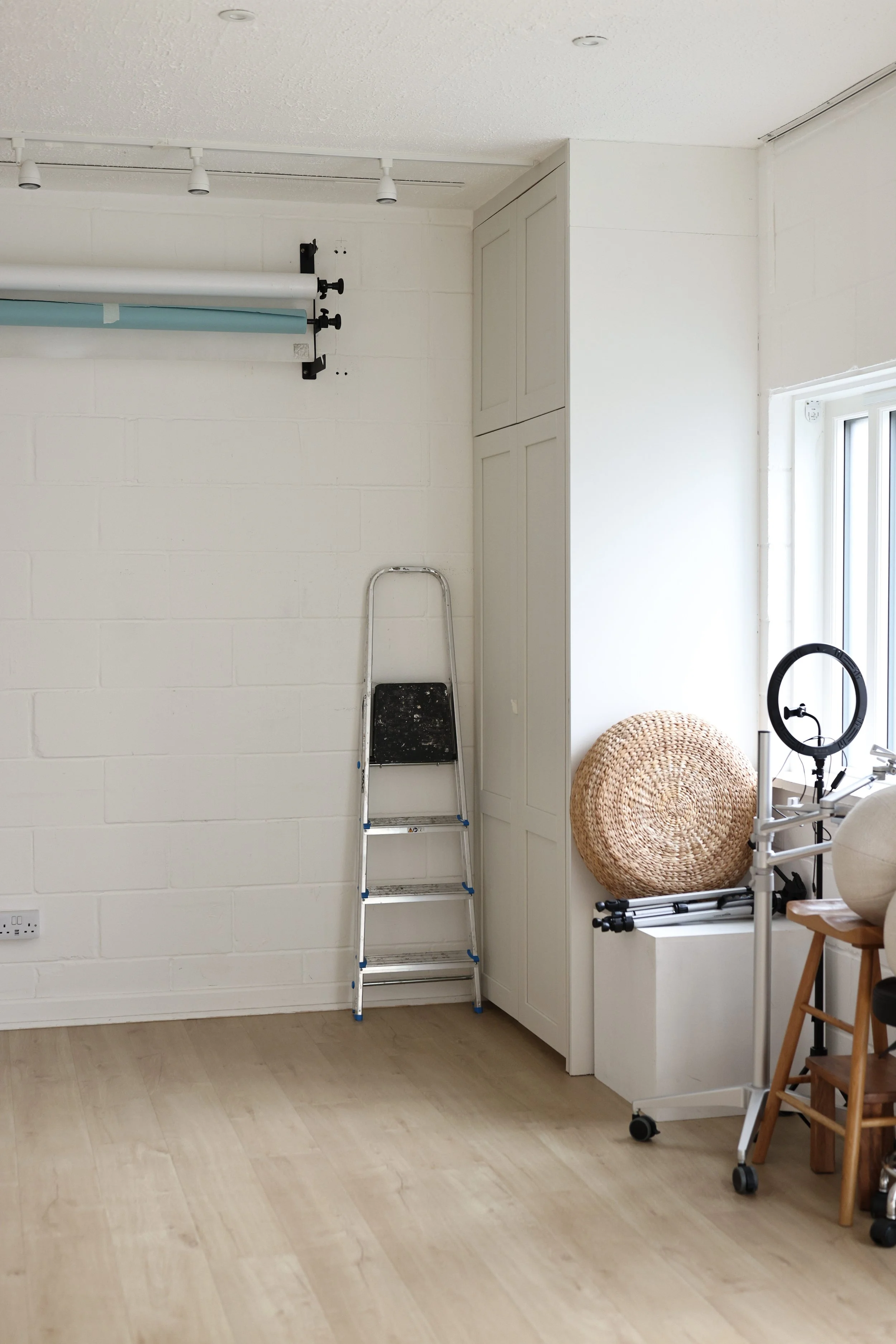 Interior of a room with white walls and light wood flooring. There is a metal step ladder, a woven round decor piece, a small table with a ring light, and a cabinet along the wall.