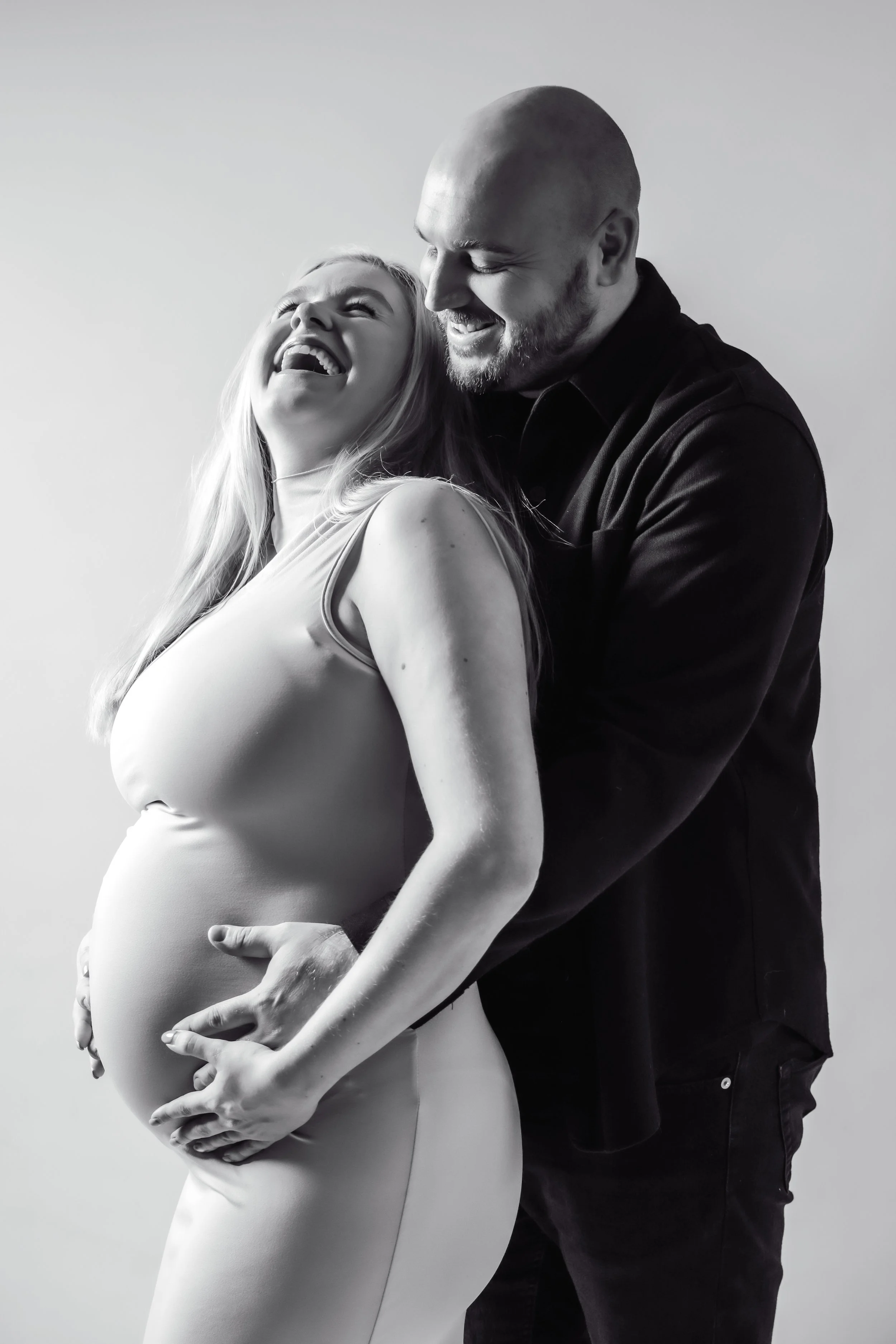 A pregnant woman and a man are laughing and embracing each other in a black and white photo.