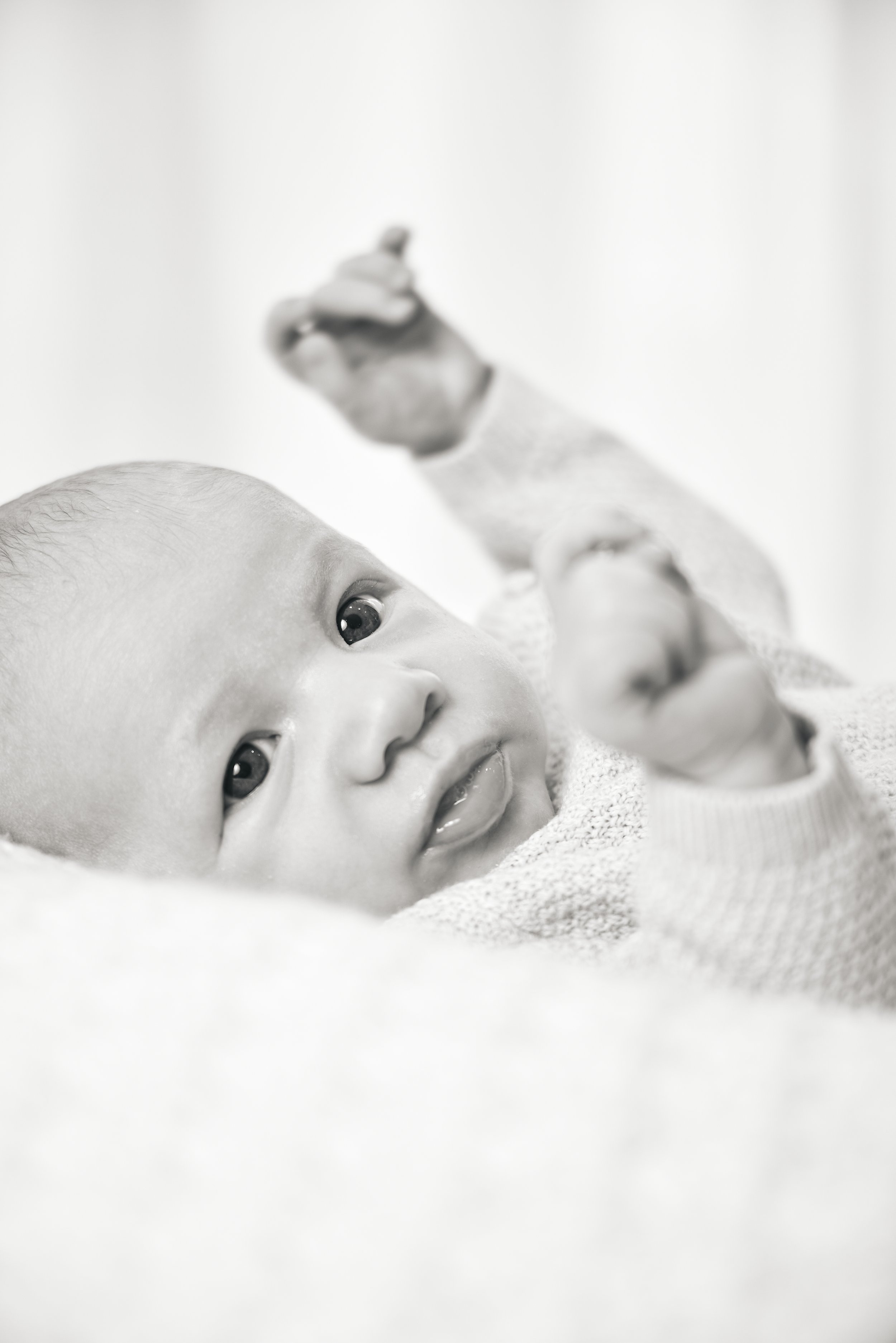 Close-up of a baby lying on a soft surface, looking at the camera with a slight smile, with one arm raised and out of focus in the background.