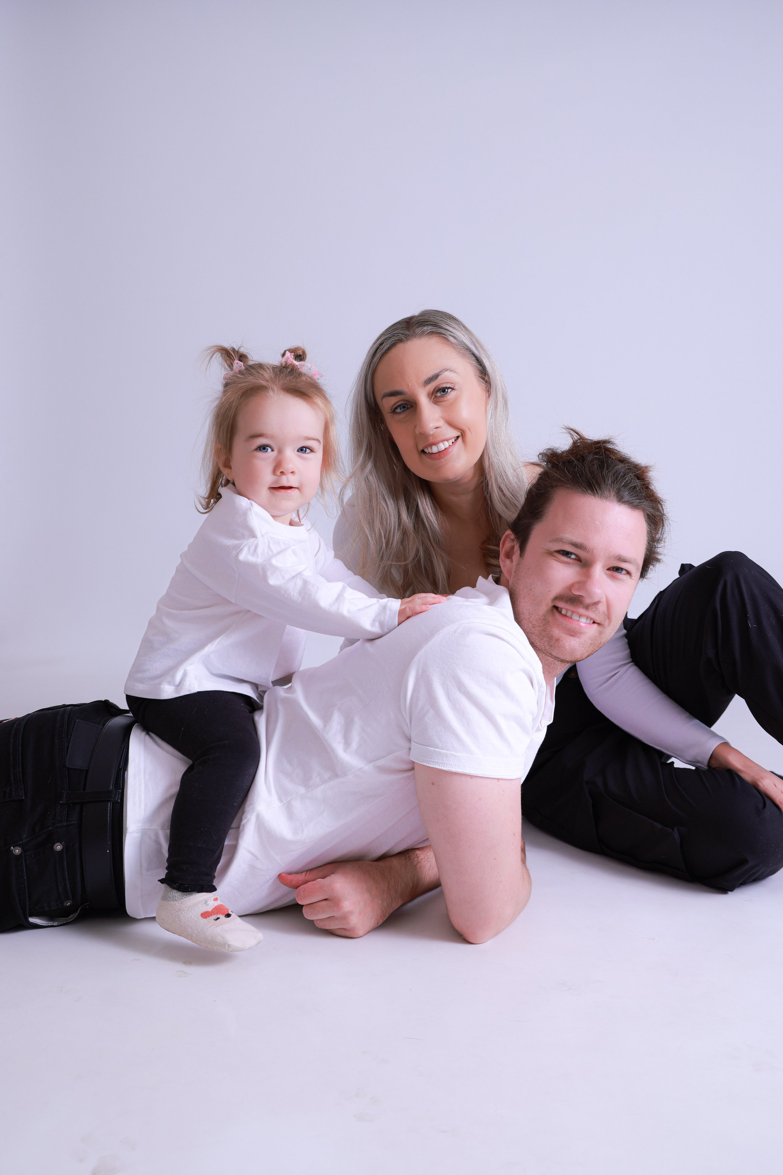 A family of three sitting and lying on the floor, smiling at the camera against a white background.
