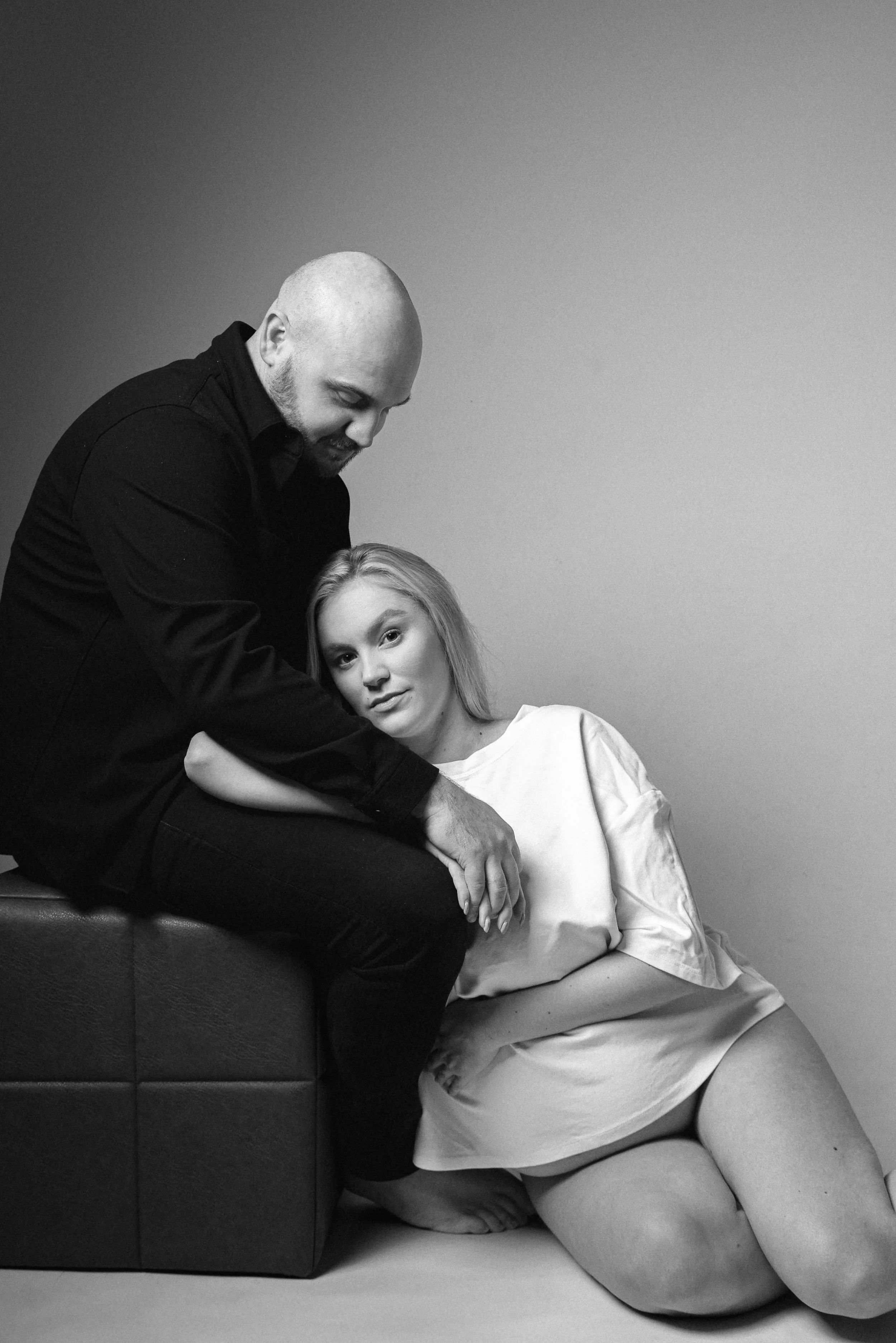 A man with a bald head and beard sitting on a cushioned surface, leaning over and holding hands with a woman with long blonde hair, who is sitting or kneeling on the floor wearing a loose white shirt, looking at the camera.