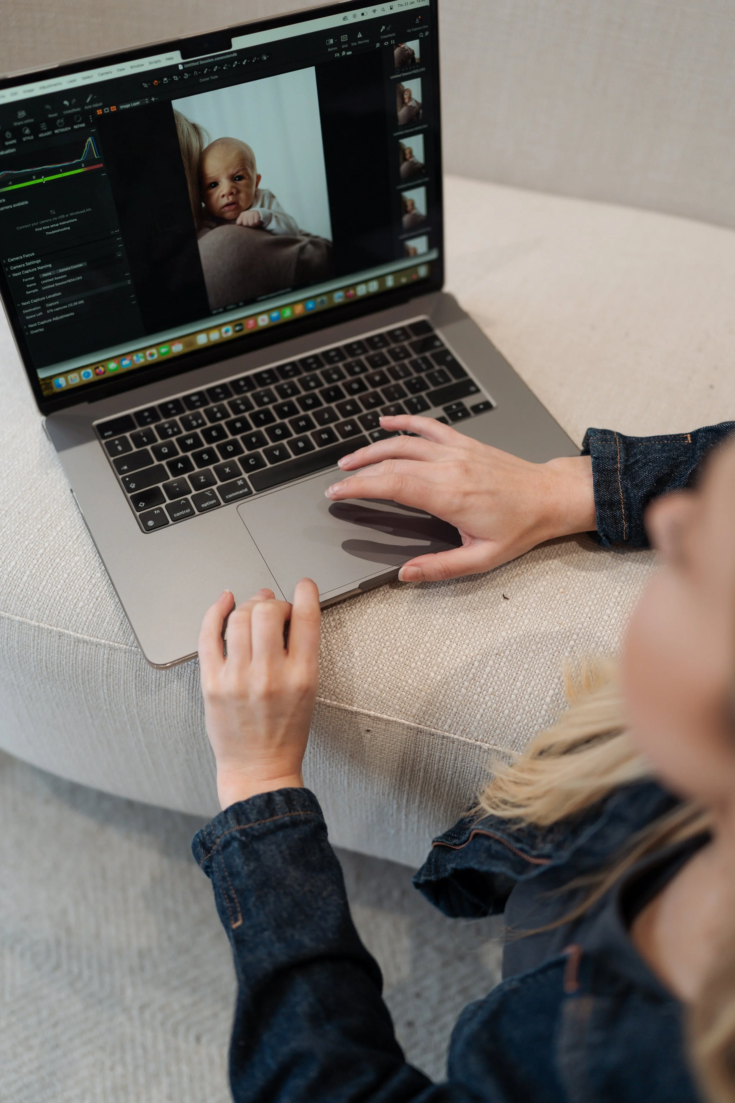A person is working on a laptop computer, editing a photo of a baby on photo editing software. The person is wearing a denim jacket and is sitting on a beige sofa.