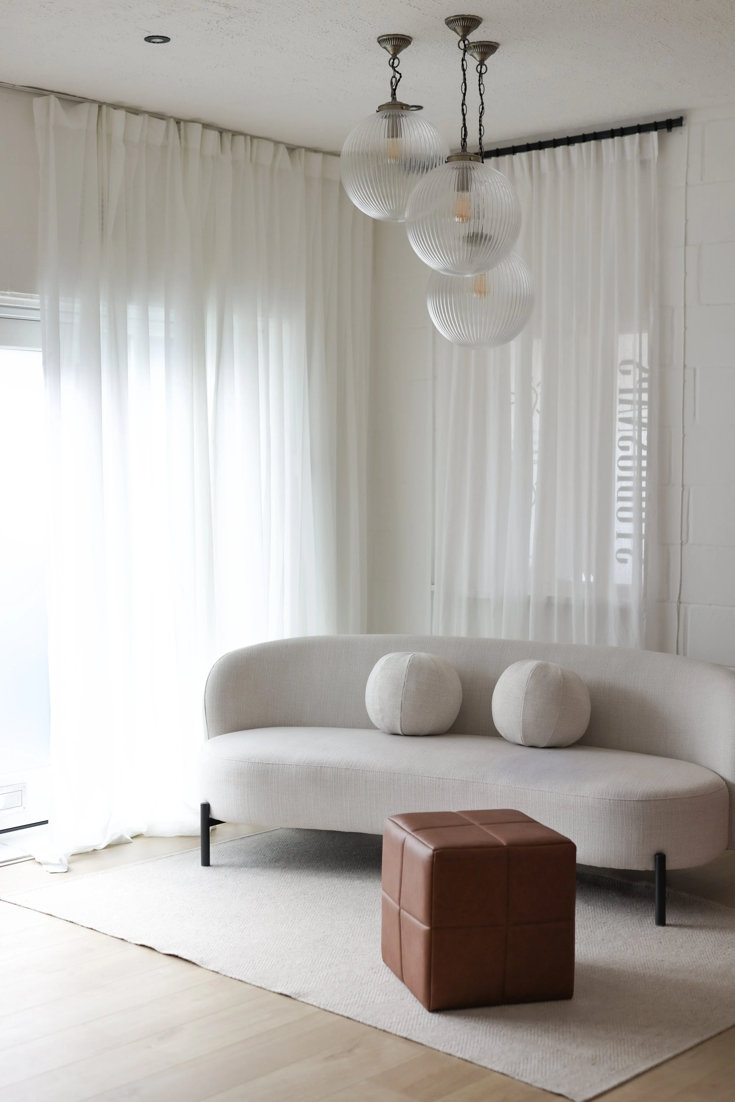 Bright living room with white curtains, minimalist white sofa, two round beige pillows, brown leather cube ottoman, hanging pendant lights with three spherical glass shades.