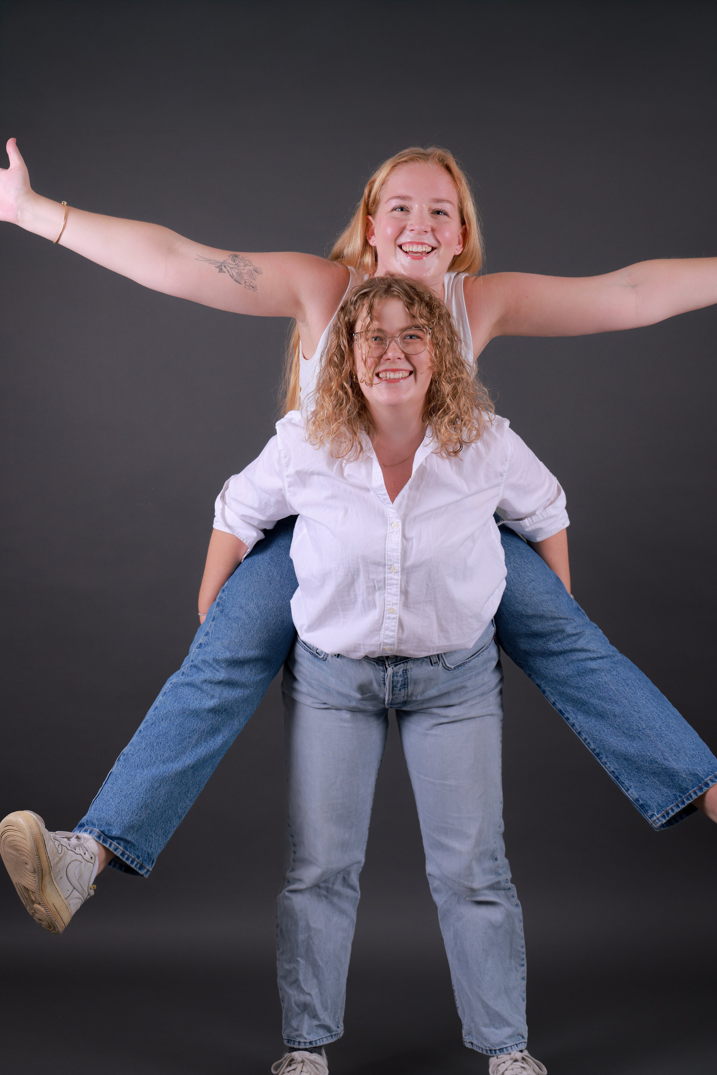 Two young women, one with red hair and one with curly brown hair and glasses, playfully stacked on a dark gray background, smiling and expressing joy.