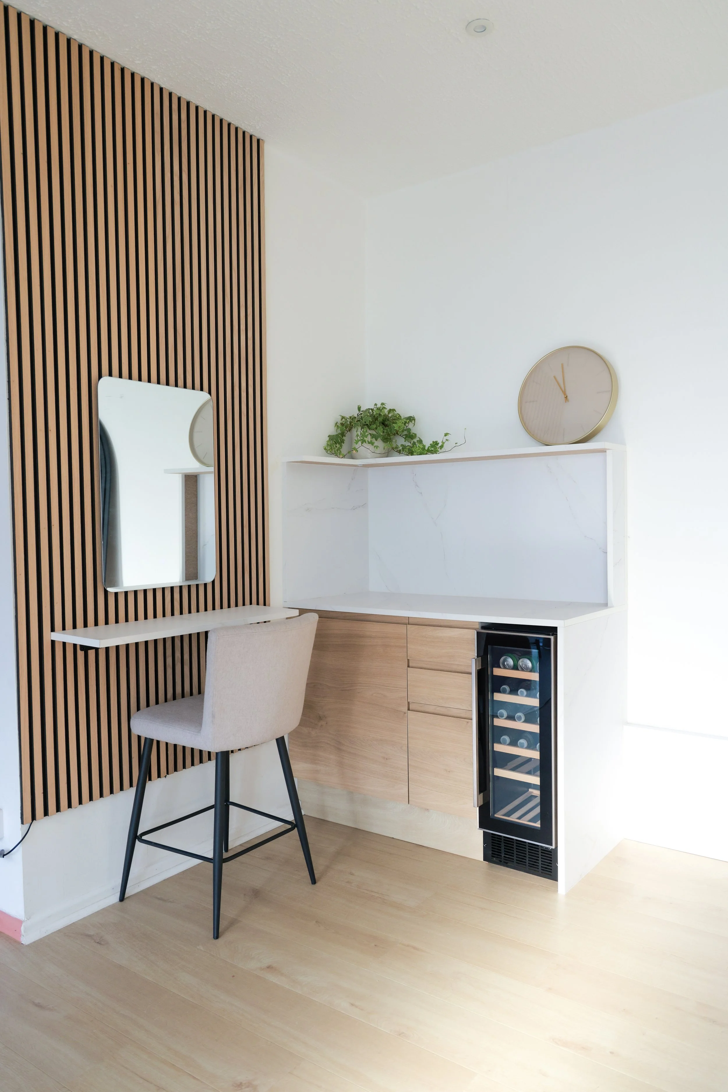 A modern minimalist interior space with a small desk, beige upholstered chair, a mirror on a slatted wooden wall, a white marble countertop with drawers, a small wine cooler, a plant, and a round wall clock.