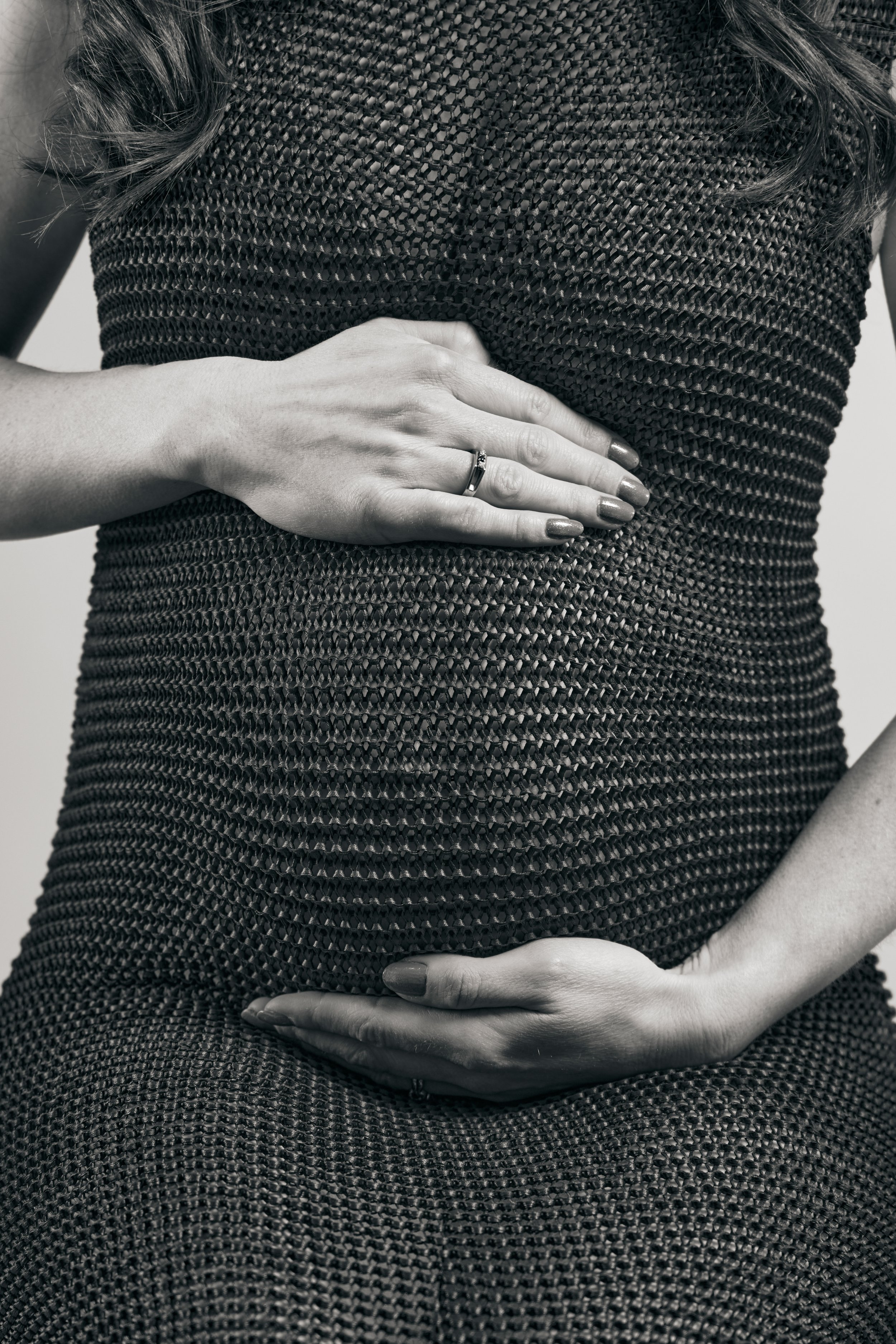 A close-up black and white photo of a woman's midsection, with one hand on her stomach and the other on her side. She is wearing a textured, woven dress and has long hair.