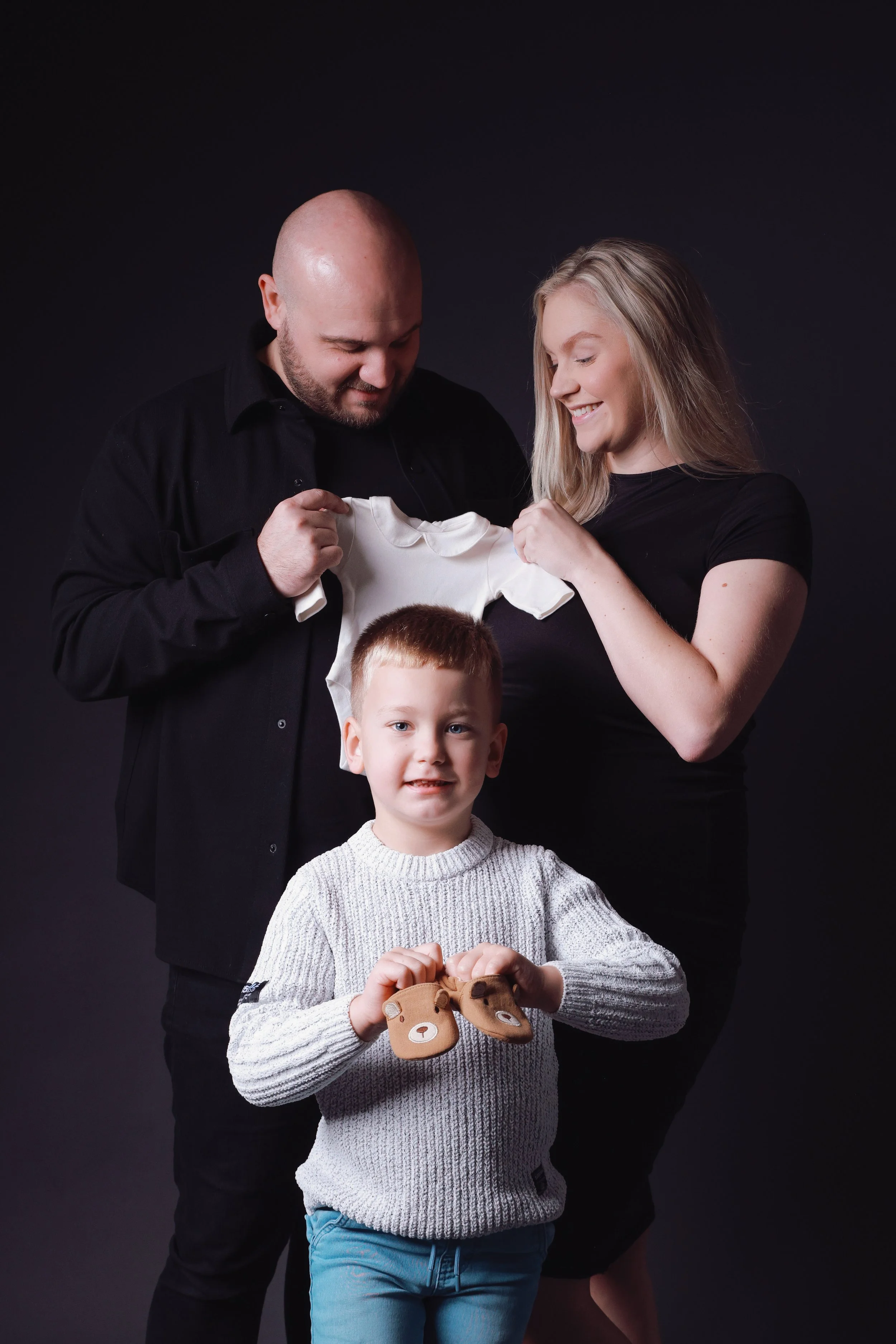 A family of three stands against a dark background. The father and mother hold a small white shirt together, smiling at it. The young boy in front holds two small bear-themed objects and looks directly at the camera.