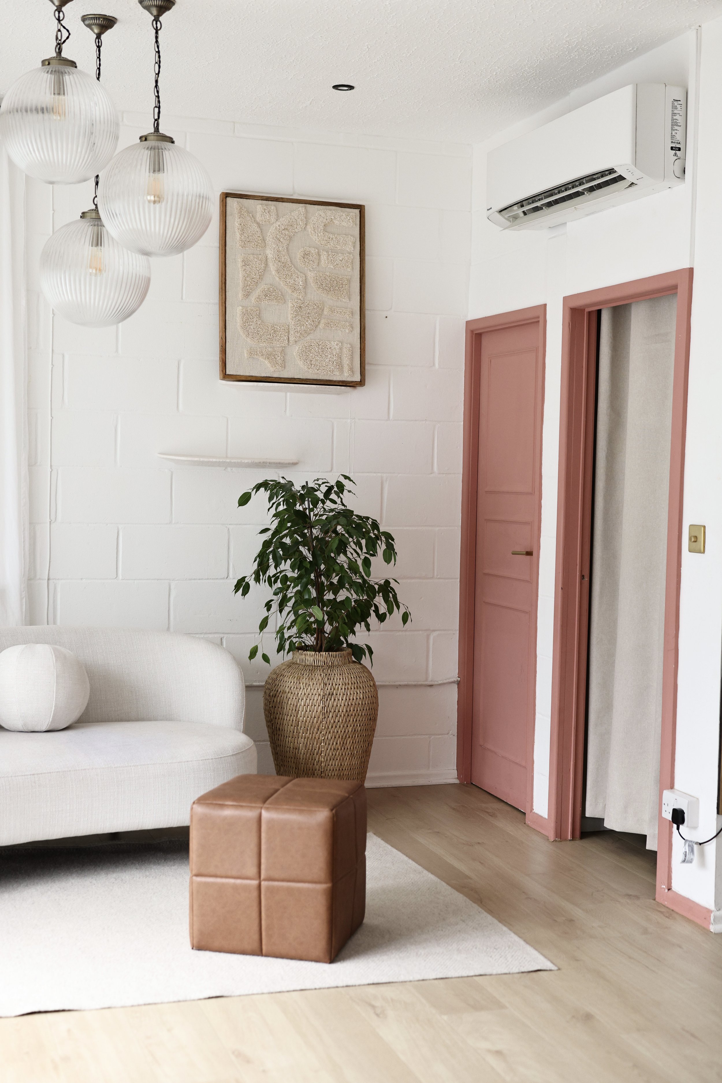 Living room with a white sofa, a tall potted plant in a woven basket, a small brown leather ottoman, pink door frames, a white wall-mounted air conditioner, and a modern light fixture with three hanging globes.