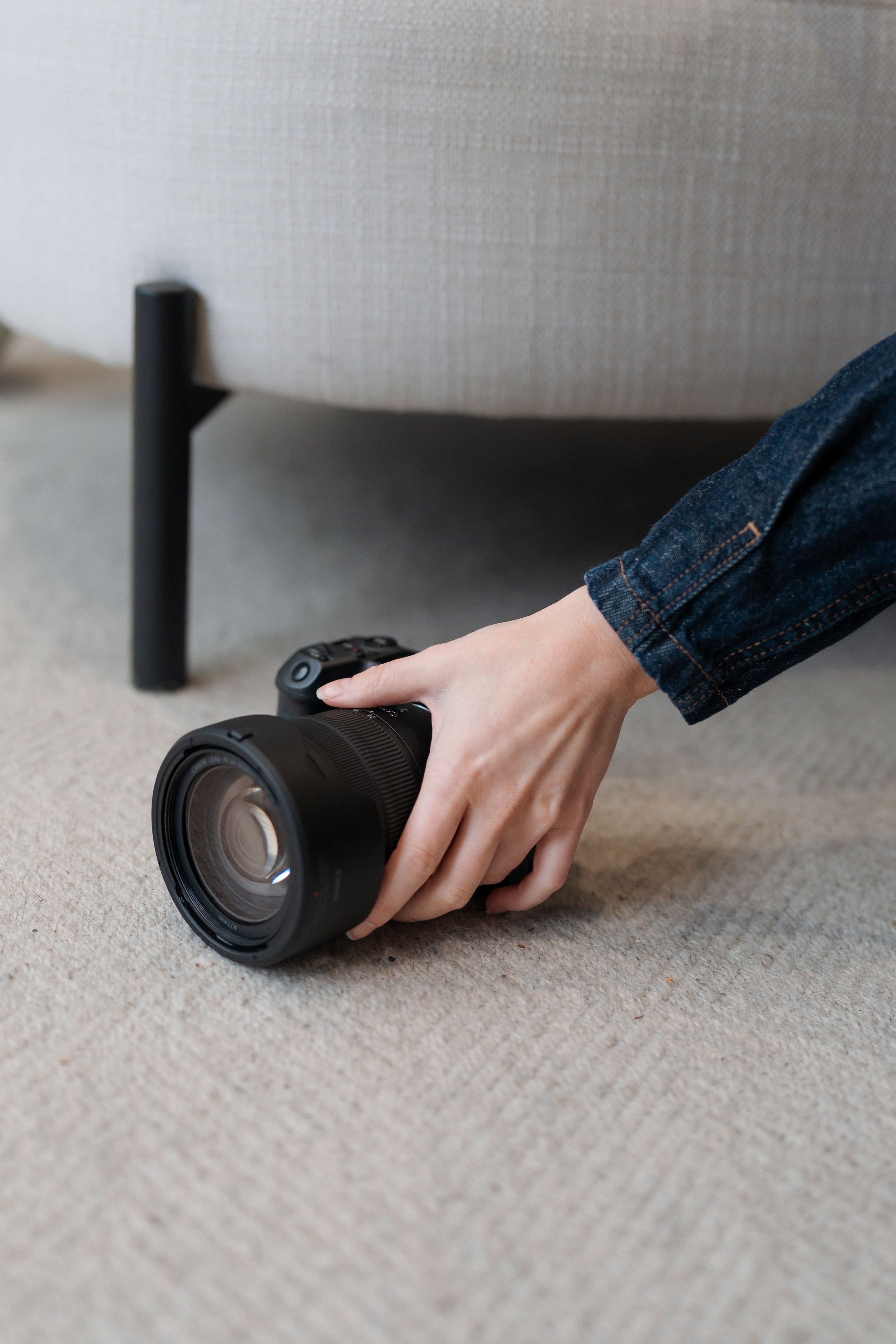 A person in a blue denim jacket is grabbing a black digital camera from the beige carpeted floor underneath a beige sofa.