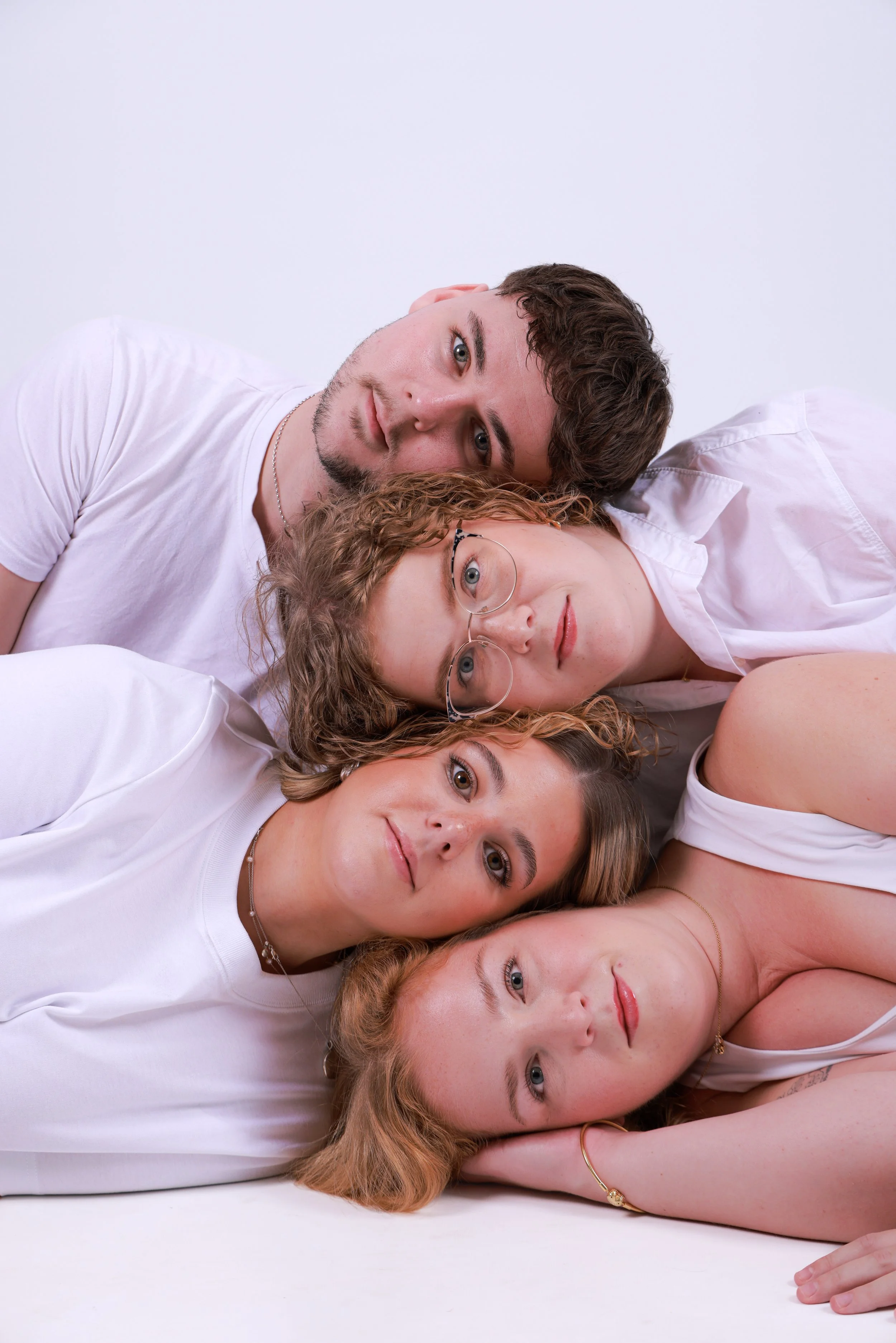 Five young adults lying close together on a white surface, with their heads touching and facing the camera.