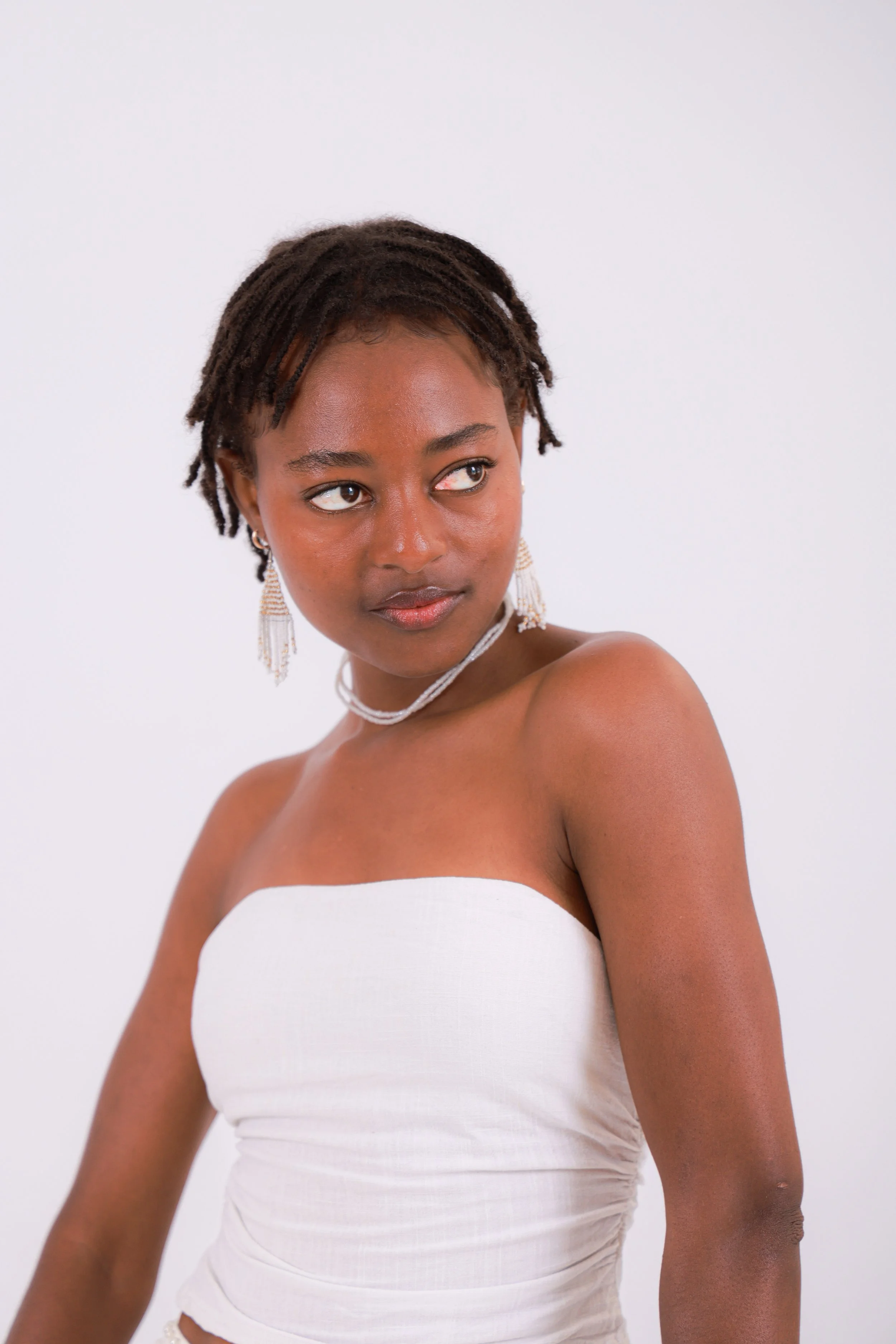 A young woman with short dreadlocks wearing a strapless white top, earrings, and a necklace, looking to her left against a plain white background.