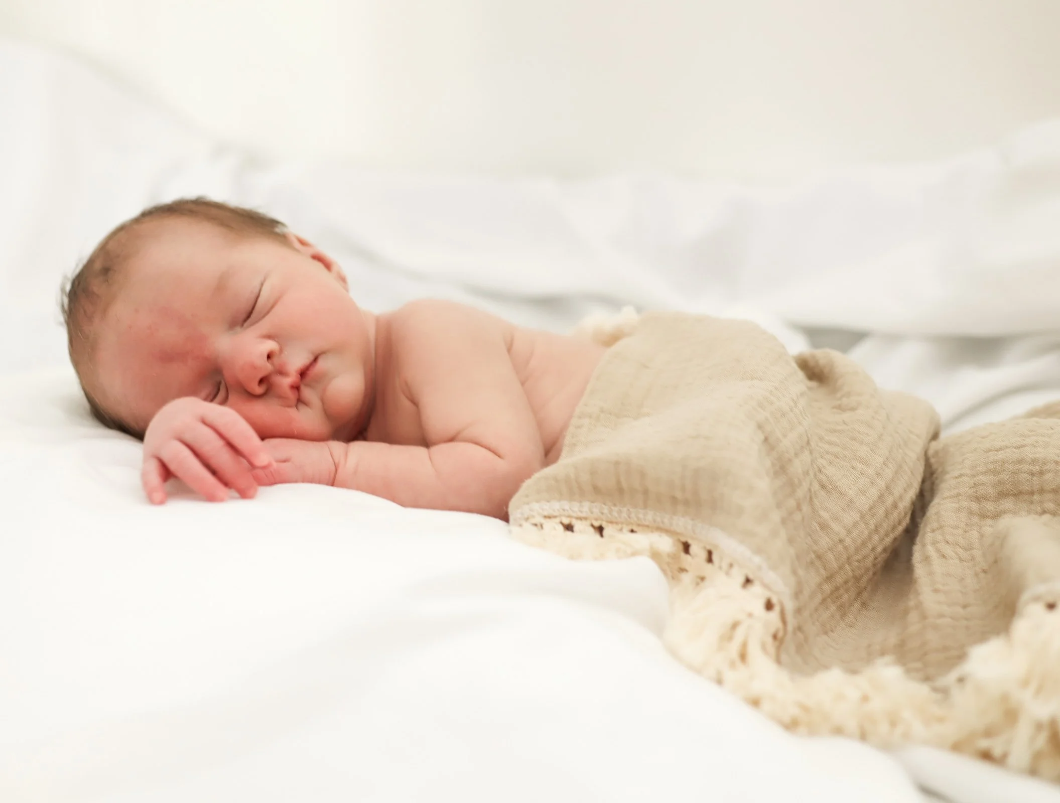 A newborn baby sleeps peacefully on a white bed, partially covered with beige knitted blanket.