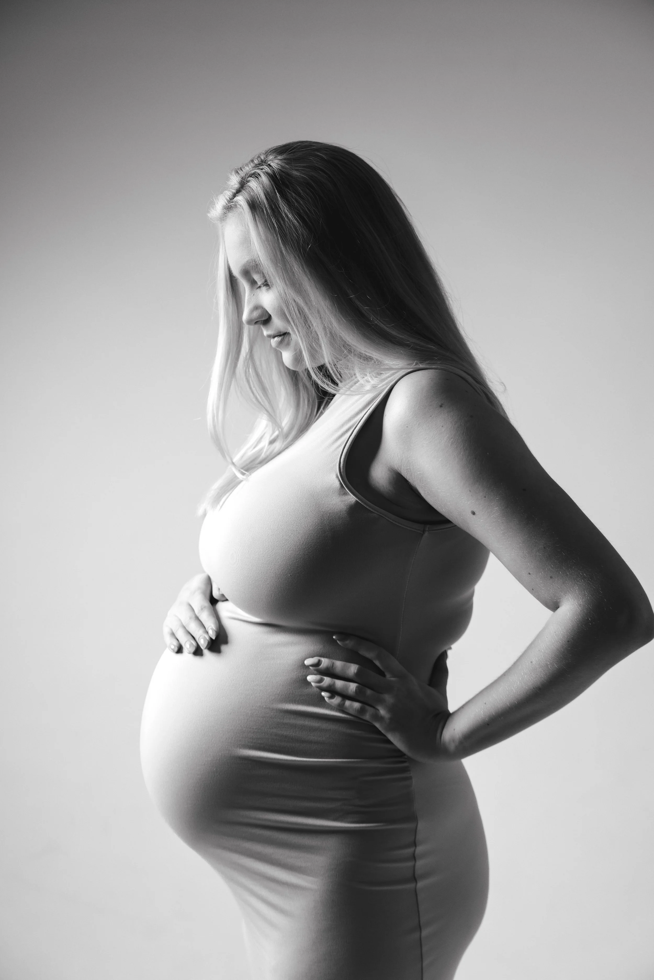 Black and white photo of a pregnant woman in a sleeveless dress, standing sideways with her hands on her belly, looking down