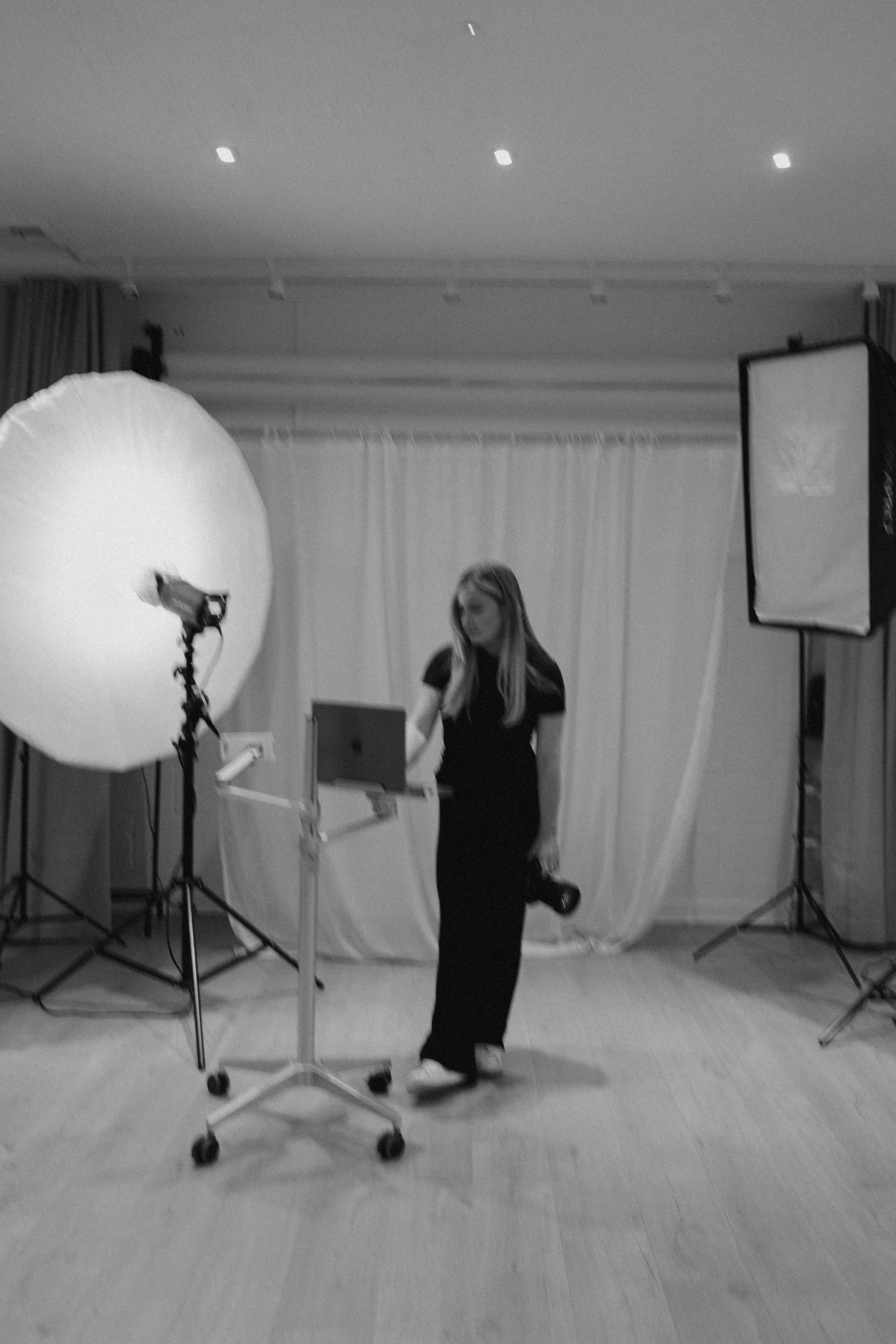 A woman in black clothes working in a photography studio with professional lighting equipment, a backdrop, and a laptop.