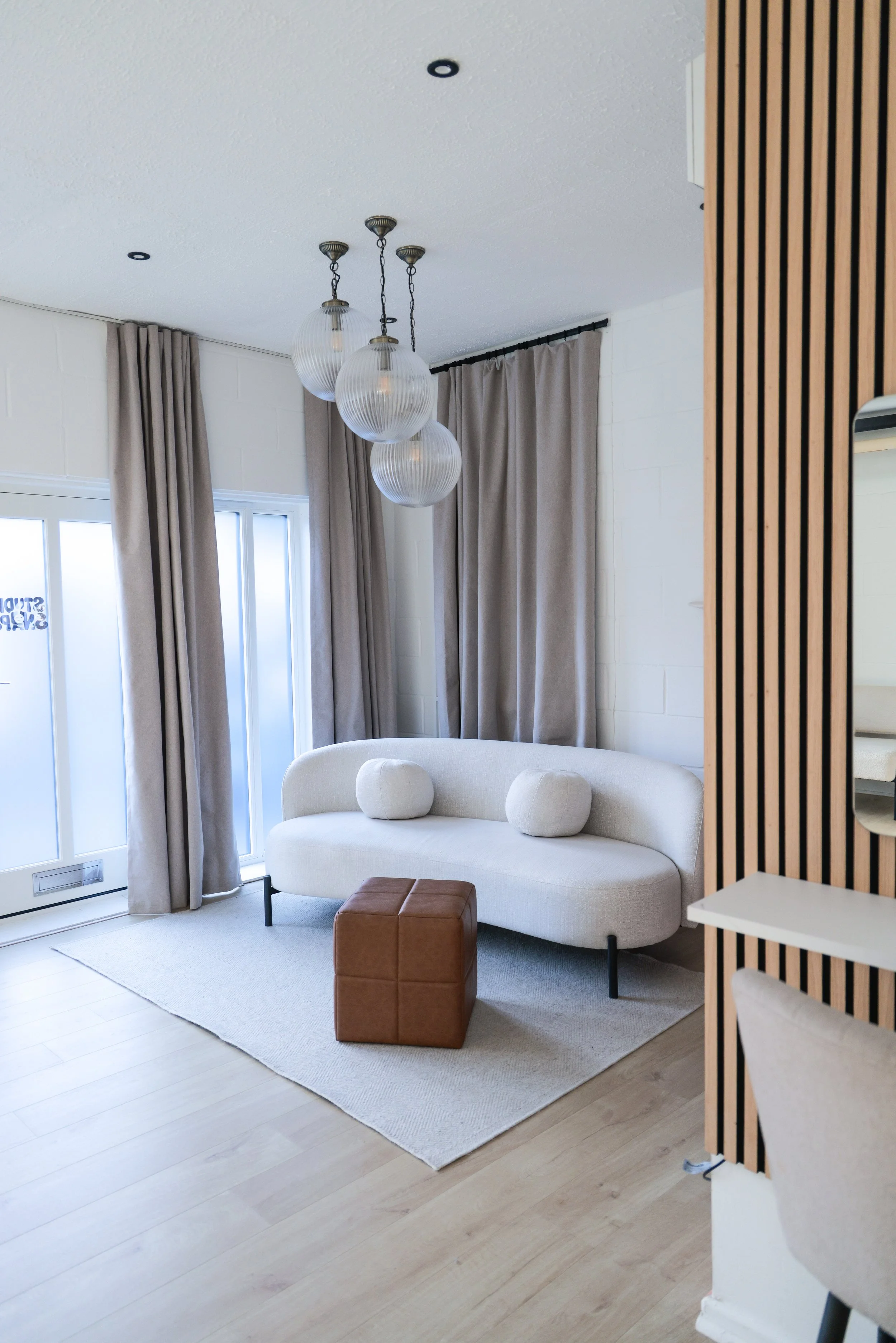 Living room with beige curtains, a white sofa with rounded cushions, a brown ottoman, a beige rug, and a modern hanging light fixture with four glass globes.