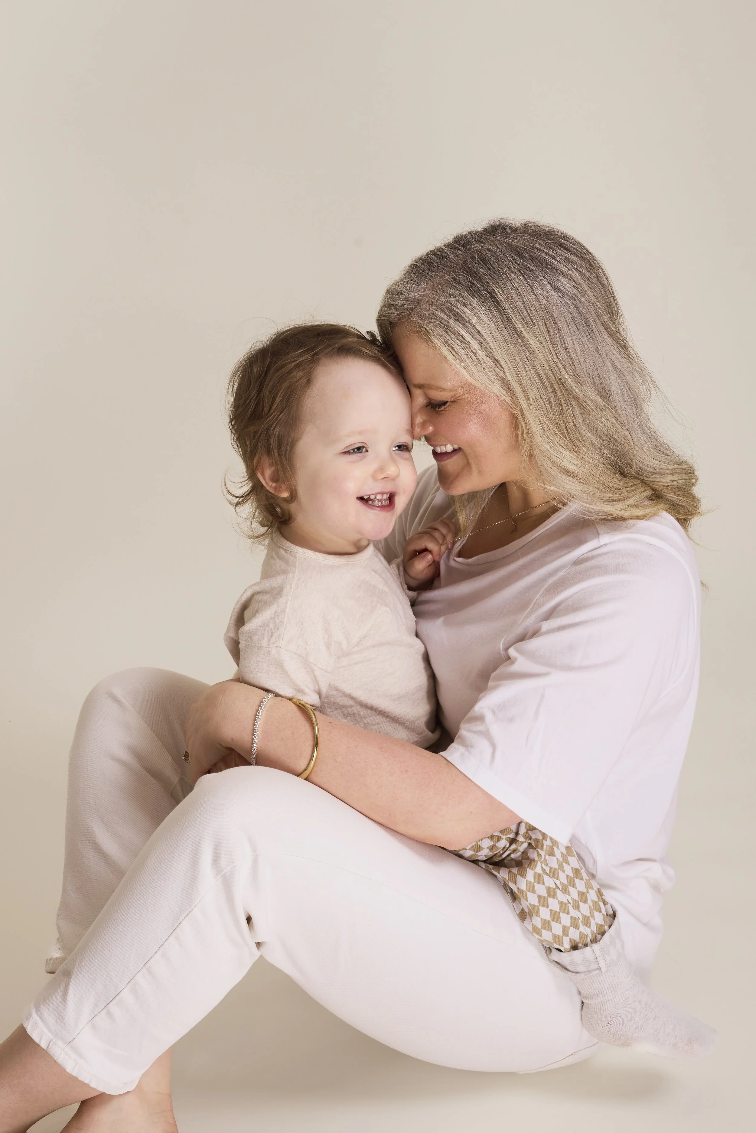 A smiling woman holding a young child close, both sharing a joyful moment with their foreheads touching against a plain beige background.