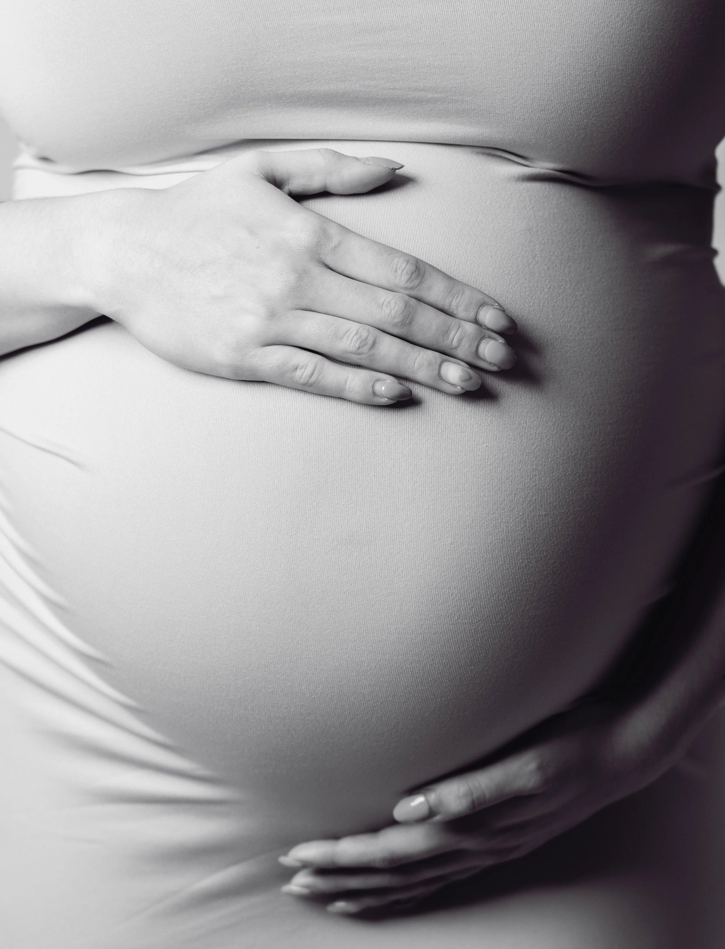 Close-up of a pregnant woman's belly with her hands gently resting on it, in black and white.