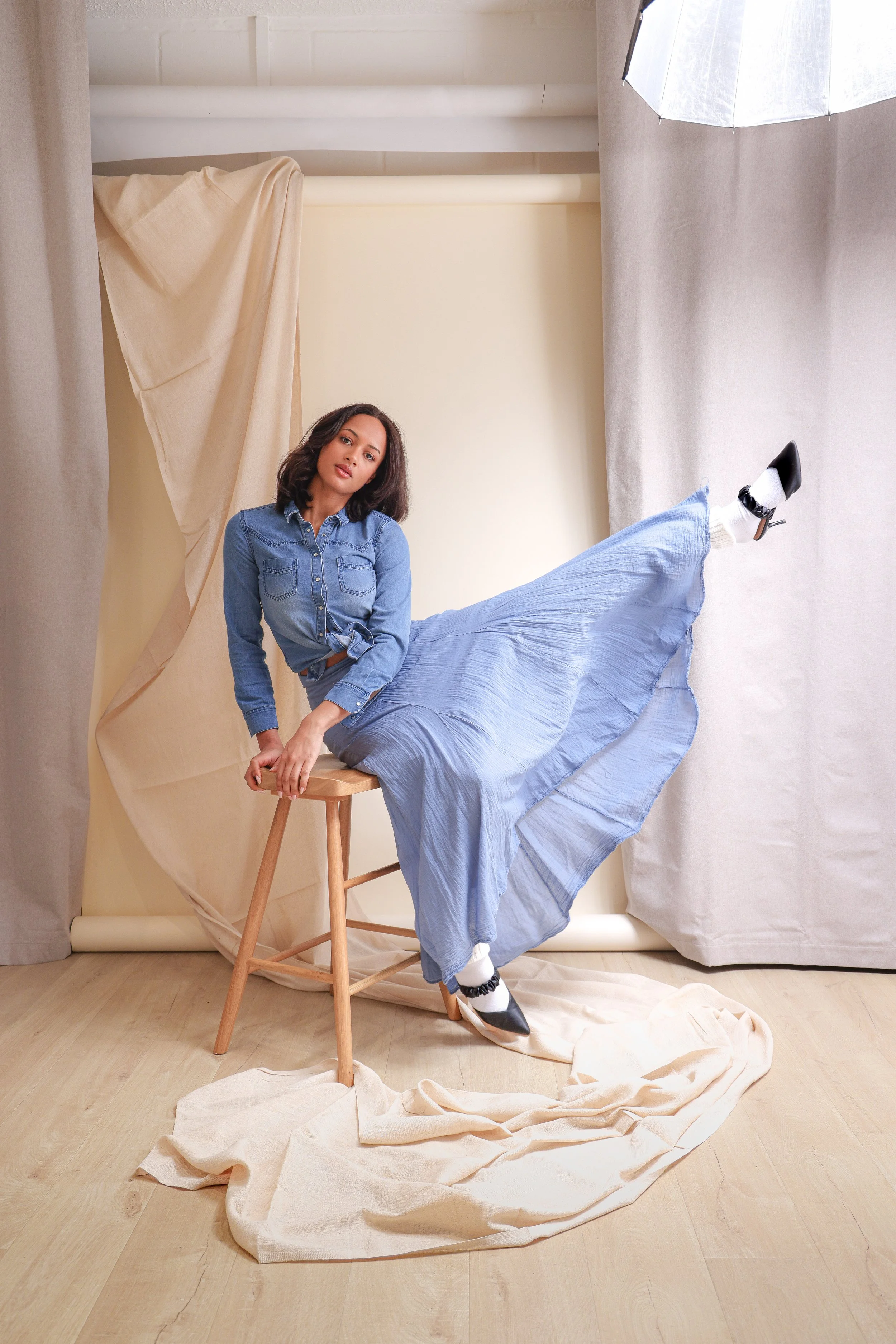 A woman in a denim shirt and blue skirt sitting on a stool with her leg extended upward, wearing black and white high heels in a studio setting with draped fabric and a soft backdrop.