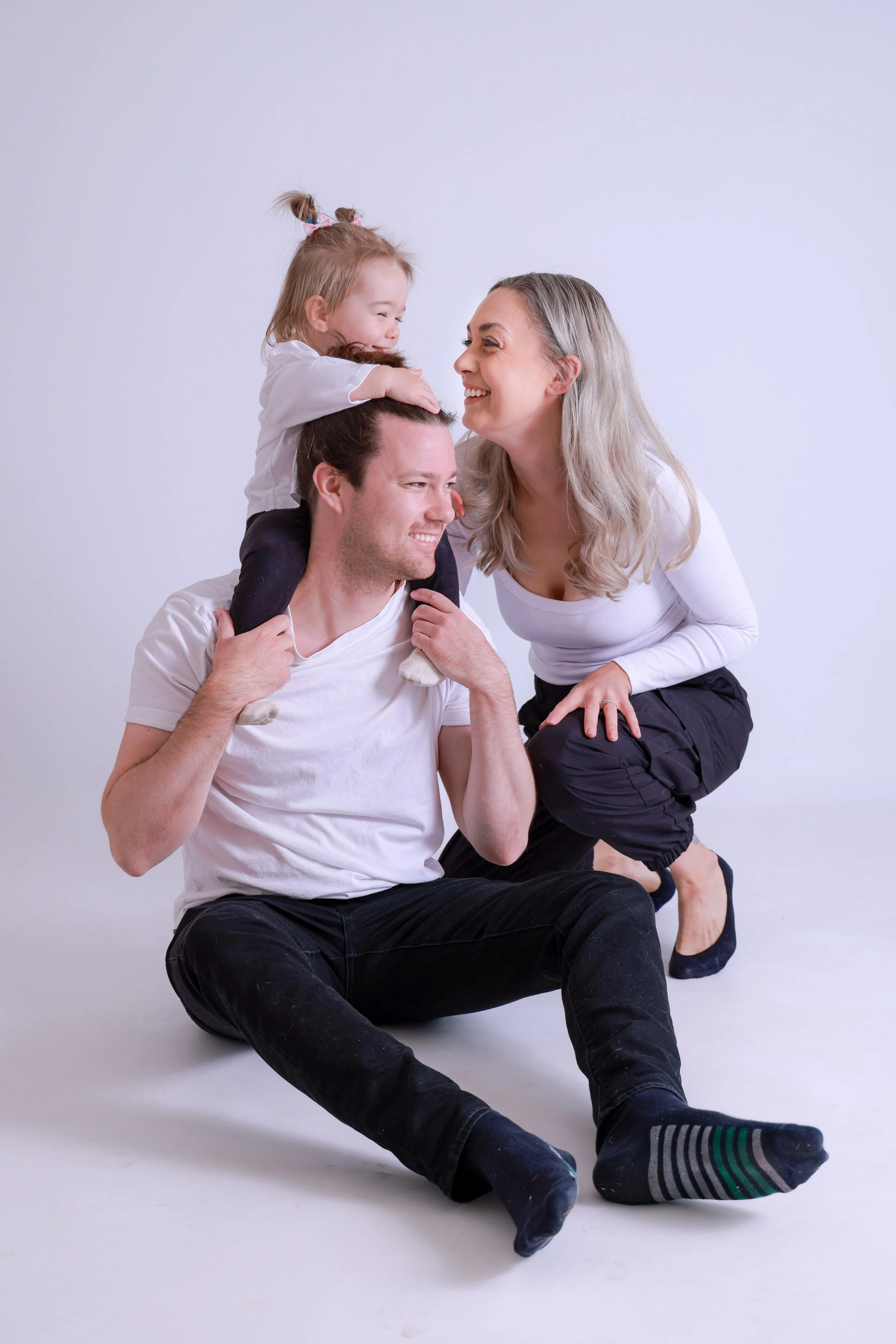 Family of three: father sitting on the floor with a young girl on his shoulders, mother crouching beside them, all smiling and interacting in a studio with a plain background.