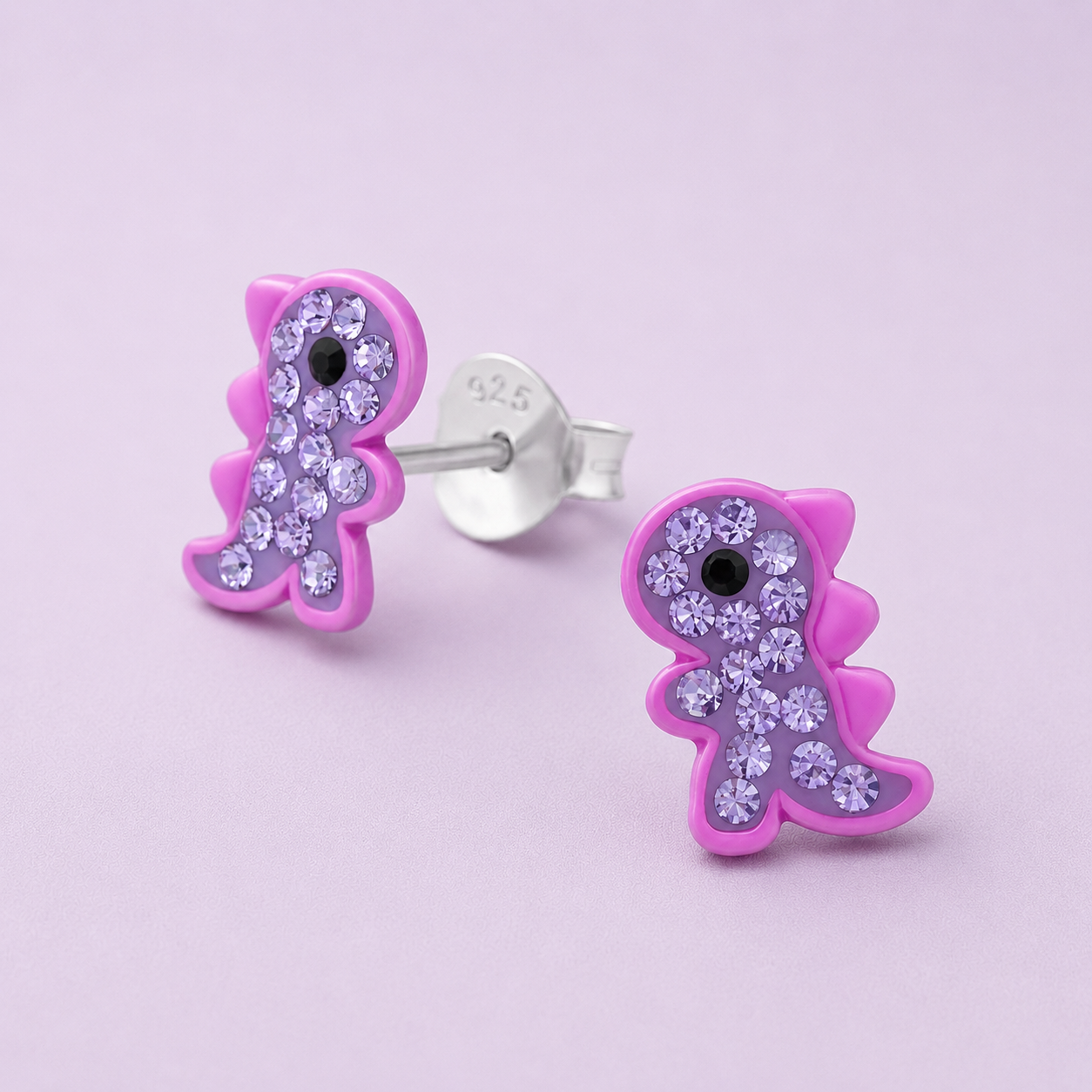 Pink dinosaur-shaped stud earrings decorated with purple rhinestones and a black center, on a pastel pink background.