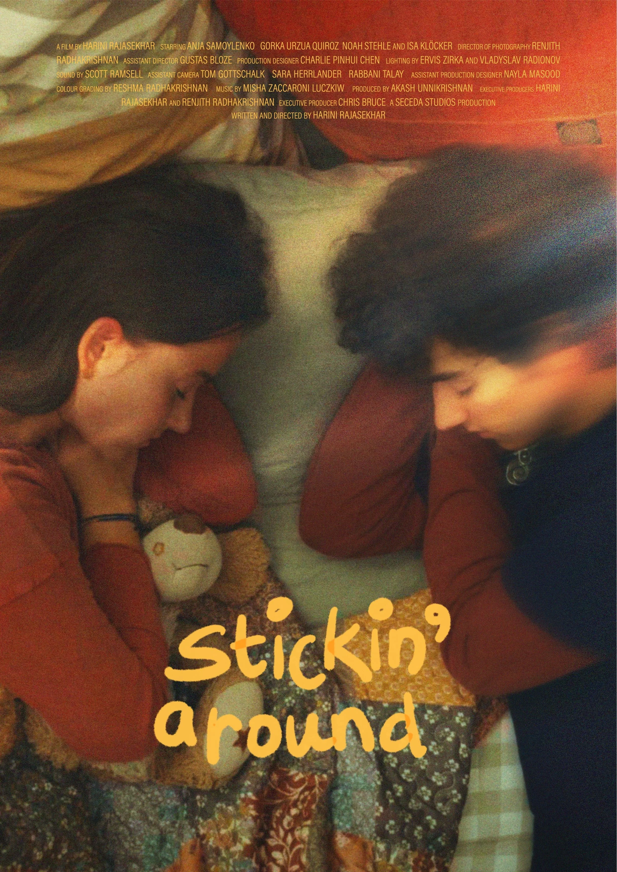 Two children with dark hair lying on their stomachs on a bed, facing each other with their heads close, surrounded by colorful blankets, with a small stuffed animal between them. The text 'sticking around' is written across the image in yellow.