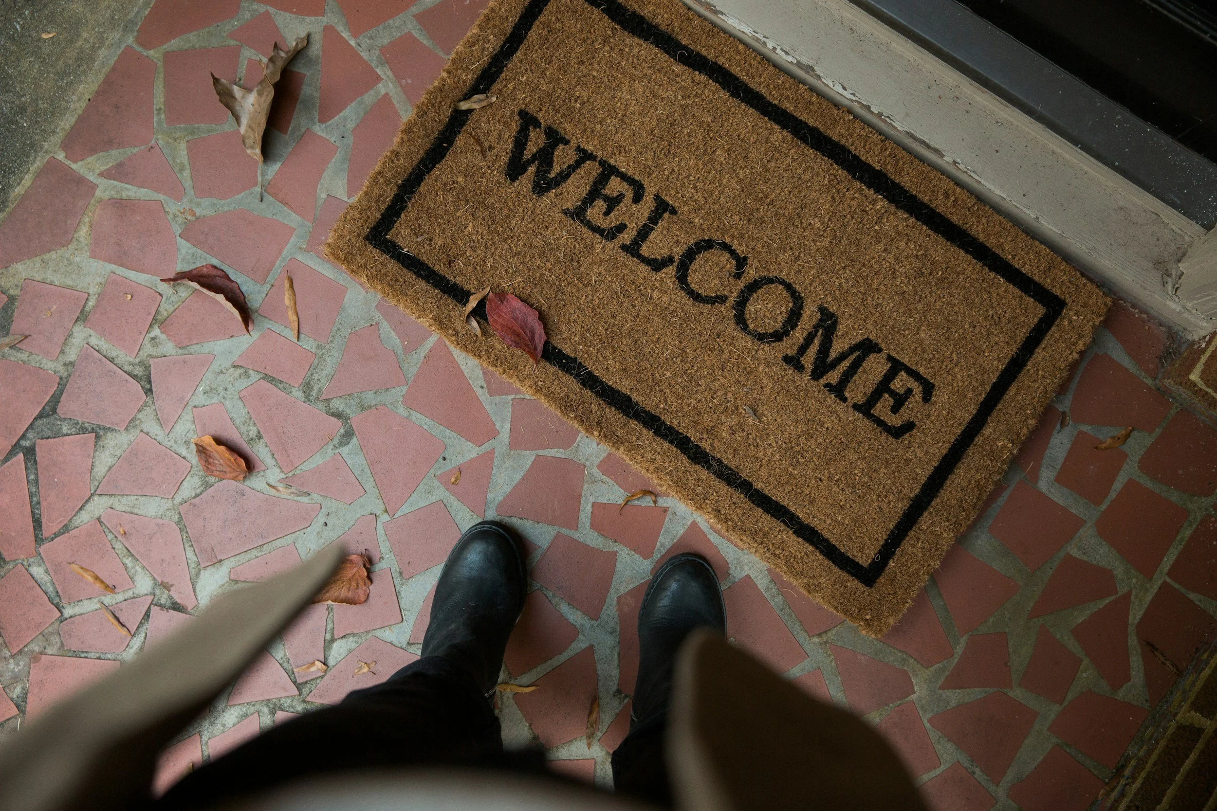 A doormat with the words 'WELCOME' printed on it, placed outside a door on a tiled porch. Some fallen leaves are scattered on the ground and on the mat, and a person's black shoes are visible in the photo.