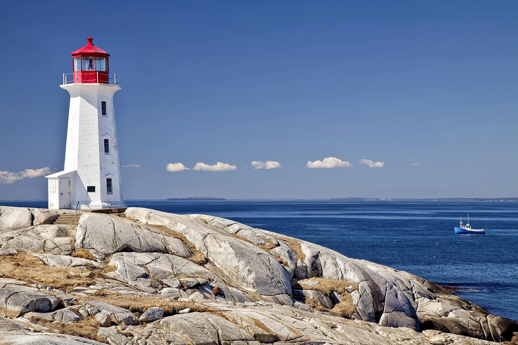 A white lighthouse with a red top on rocky terrain beside the ocean, with a boat in the water and a distant landmass under a partly cloudy sky.
