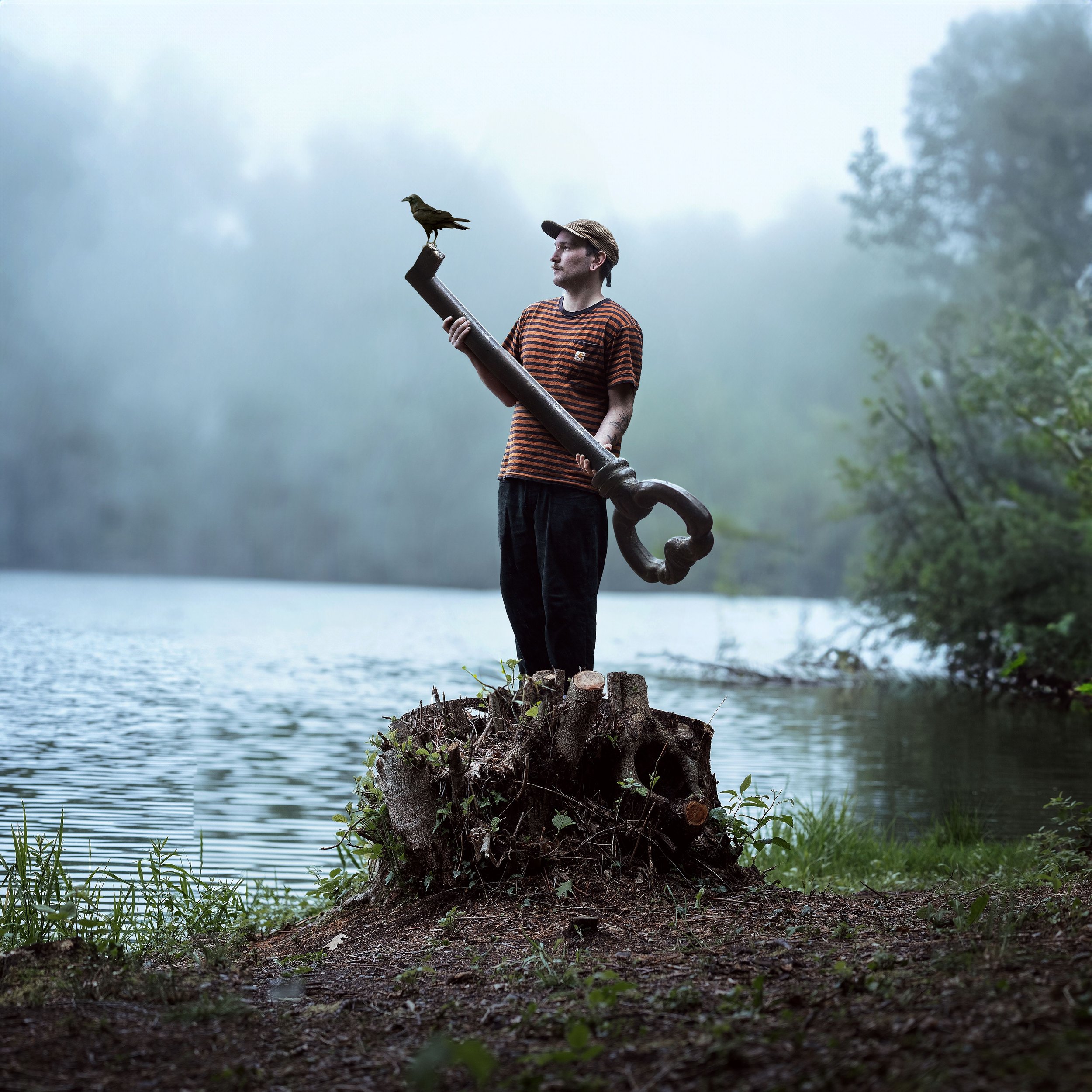 Ein junger Mann steht auf einem Baumstumpf am Seeufer und hält ein großes, altehrwürdiges großes Schlüssel-Accessoire, während eine Krähe auf dem Schlüssel sitzt. Der Hintergrund ist neblig und naturbelassen.