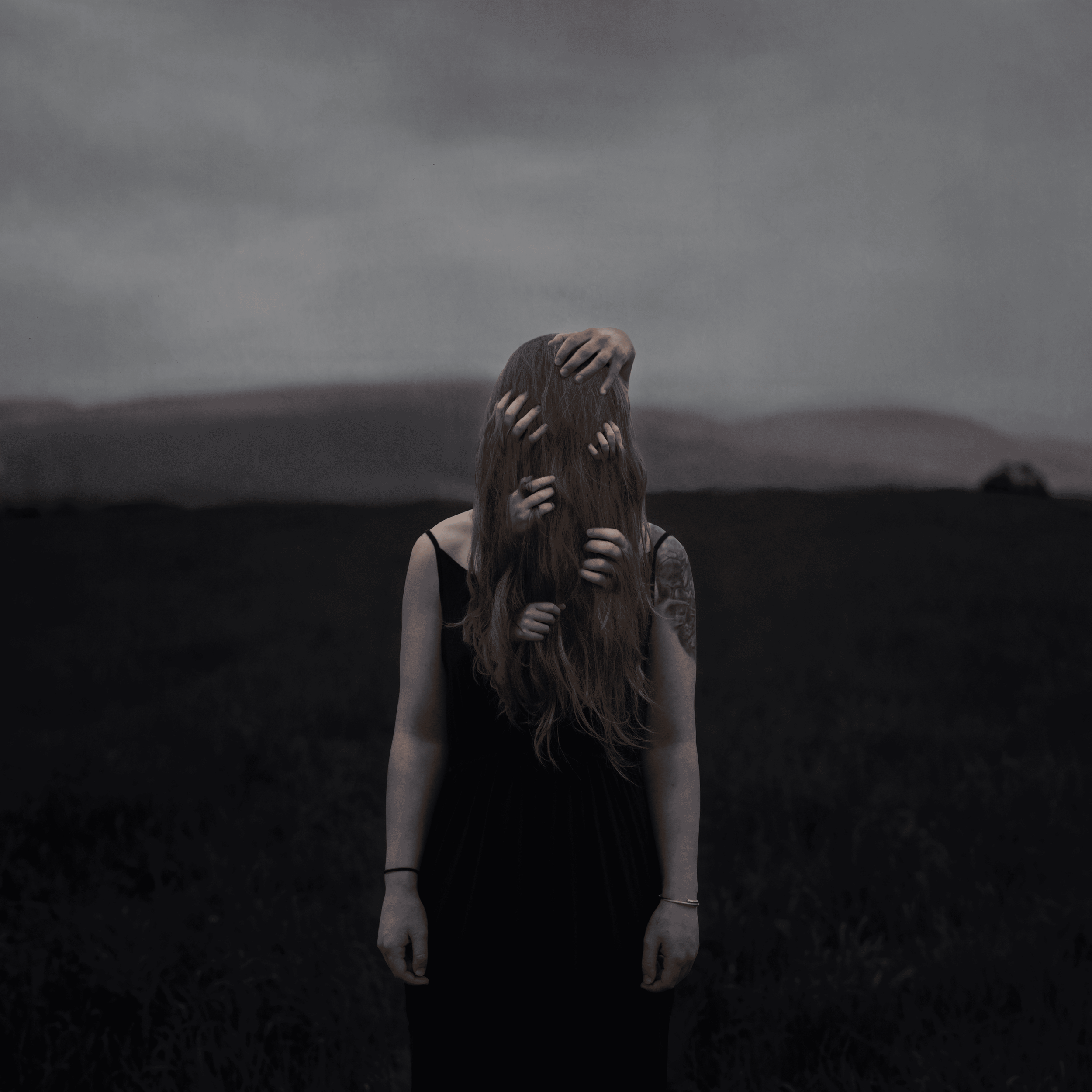 Eine Frau in einem schwarzen Kleid steht in einer dunklen Landschaft, deren Gesicht von mehreren Händen mit langen Fingern bedeckt ist.