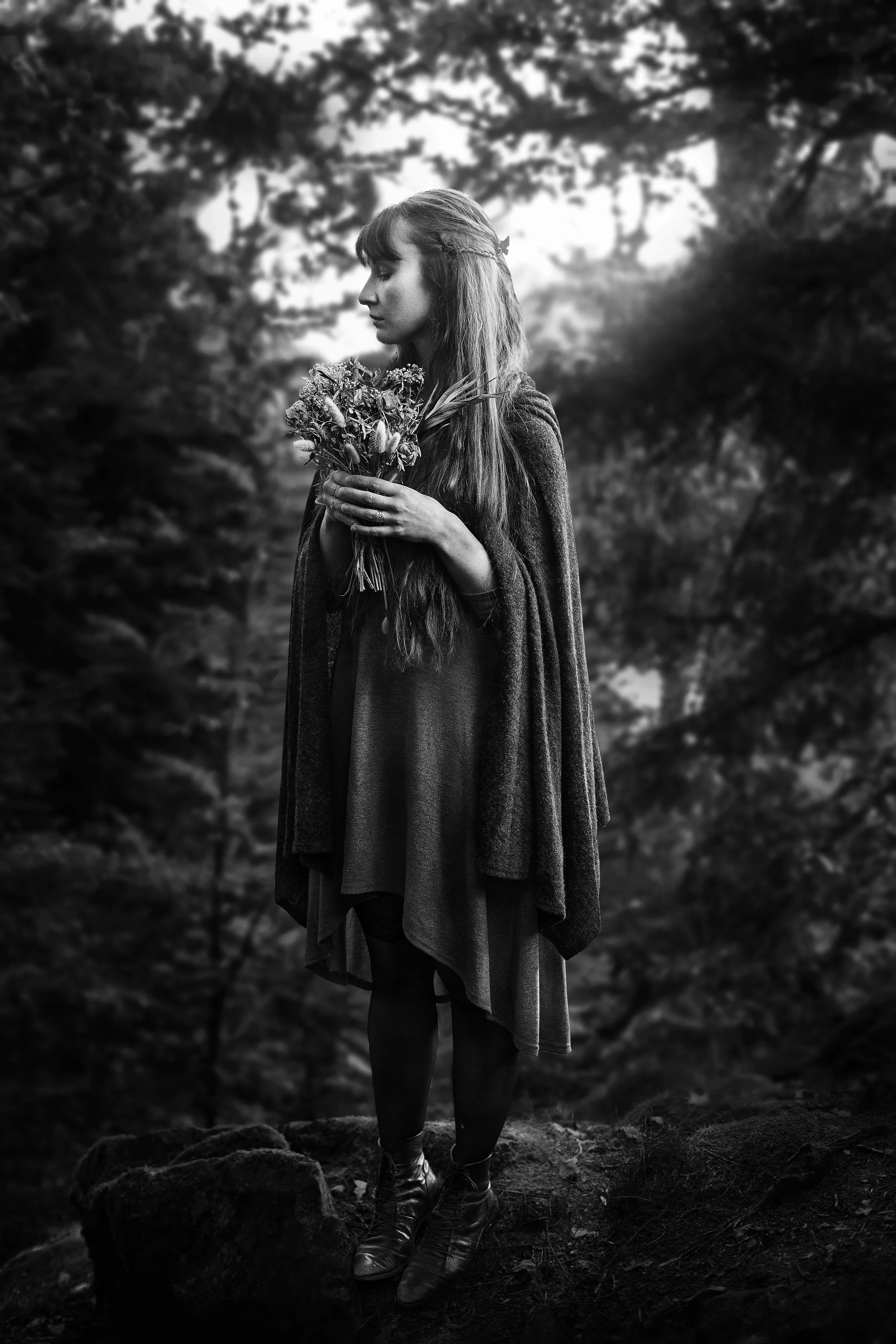 Schwarz-Weiß-Foto einer jungen Frau mit langen Haaren, die in einem dunklen Wald steht und einen Blumenstrauß in den Händen hält.