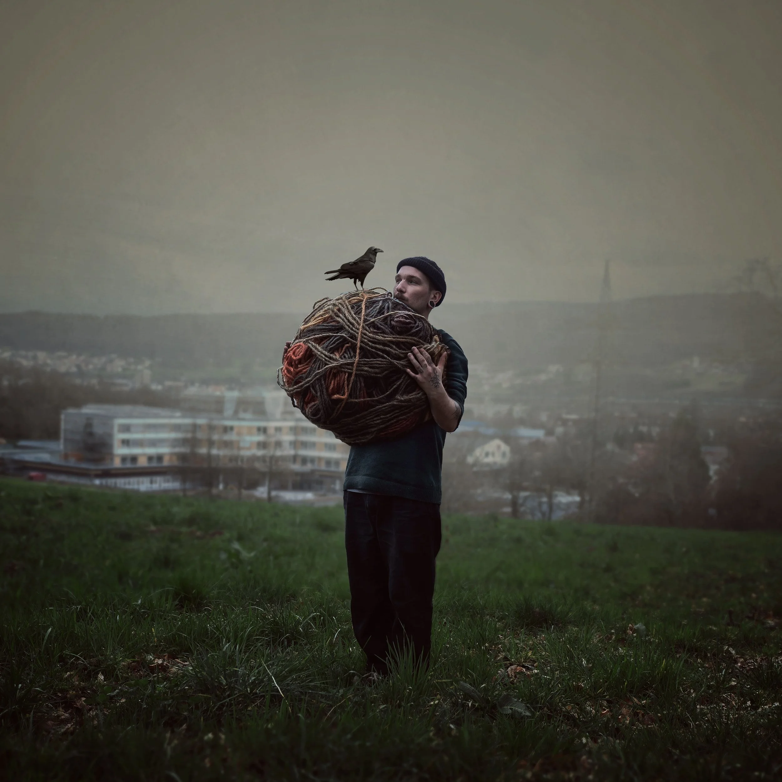 Ein junger Mann steht im Freien auf einer Wiese mit einem hohen Hügelhintergrund, trägt dunkle Kleidung und hält einen großen Wollknäuel auf seinem Schoß, auf dem ein Vogel sitzt.