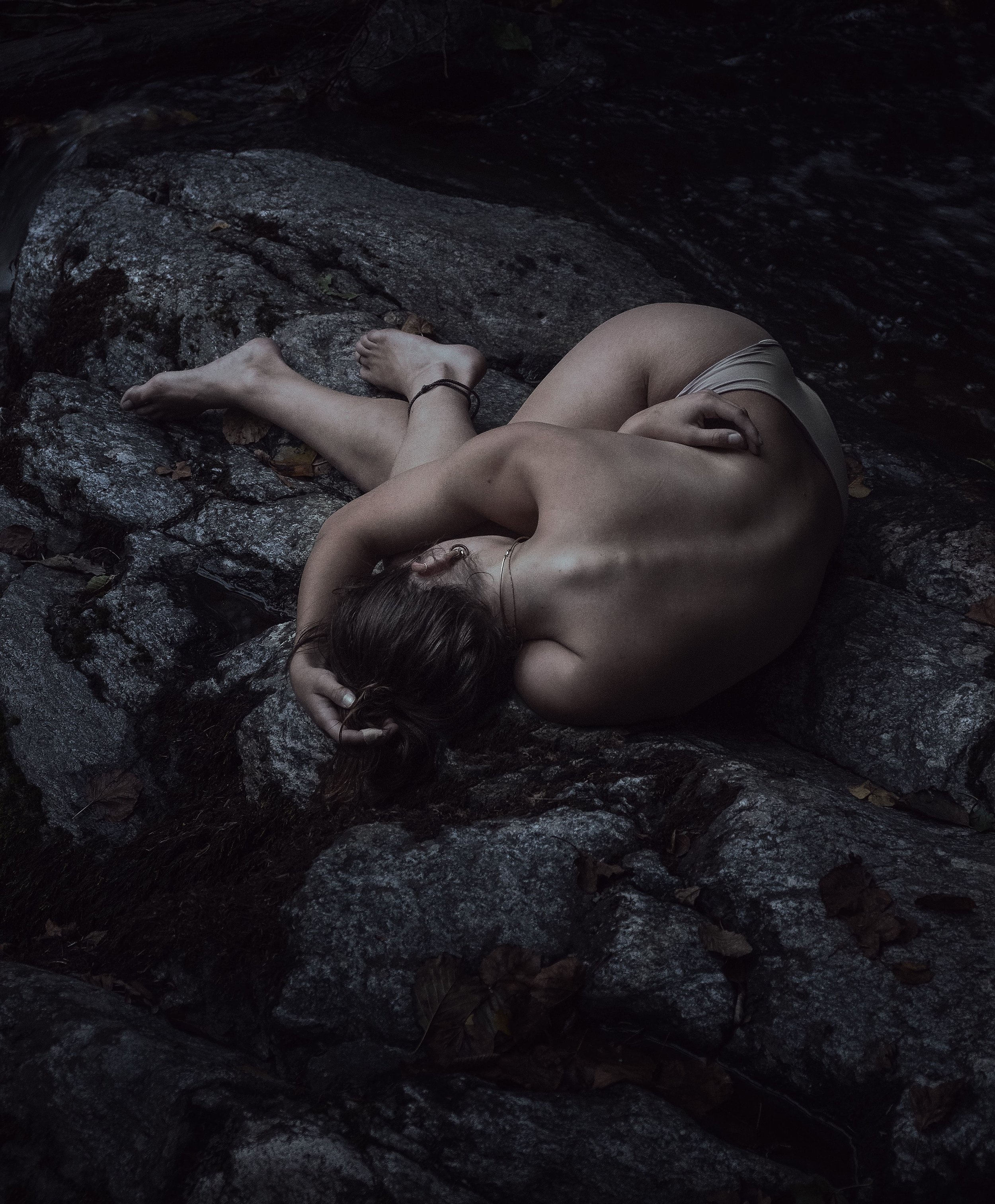 Eine nackt sitzende Frau liegt umarmt in einer felsigen, dunklen Umgebung.