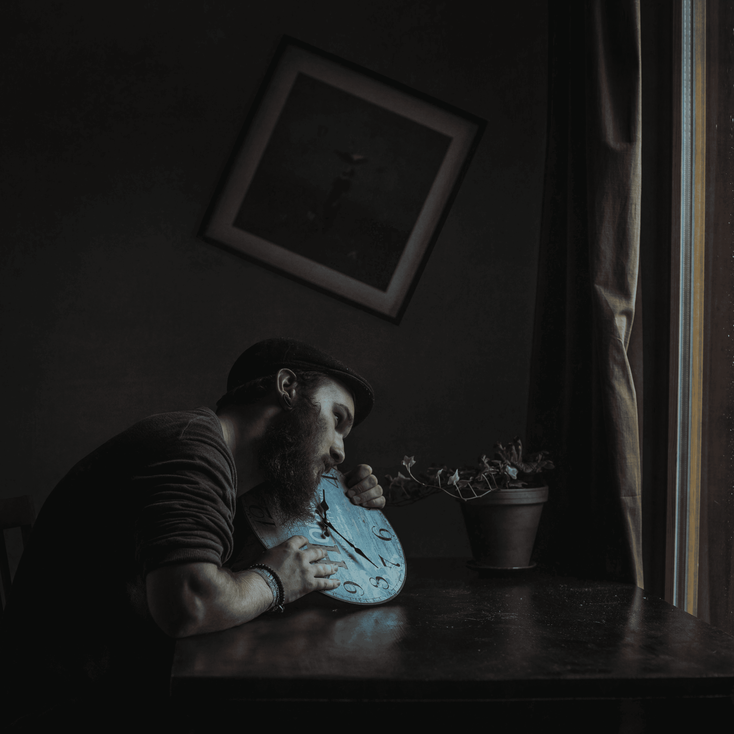 Ein Mann mit Bart und Hut, der an einem Tisch ein buntes Zifferblatt mit Zeigern umarmt, bei natürlichem Tageslicht vor einem Fenster mit Vorhang.