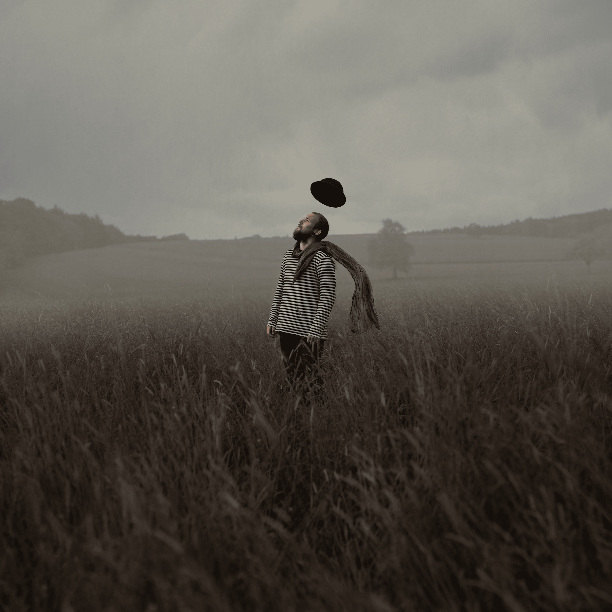 Ein Mann steht in einem Feld mit hohem Gras, trägt eine gestreifte Jacke und schaut nach oben, während ein Hut in der Luft schwebt.