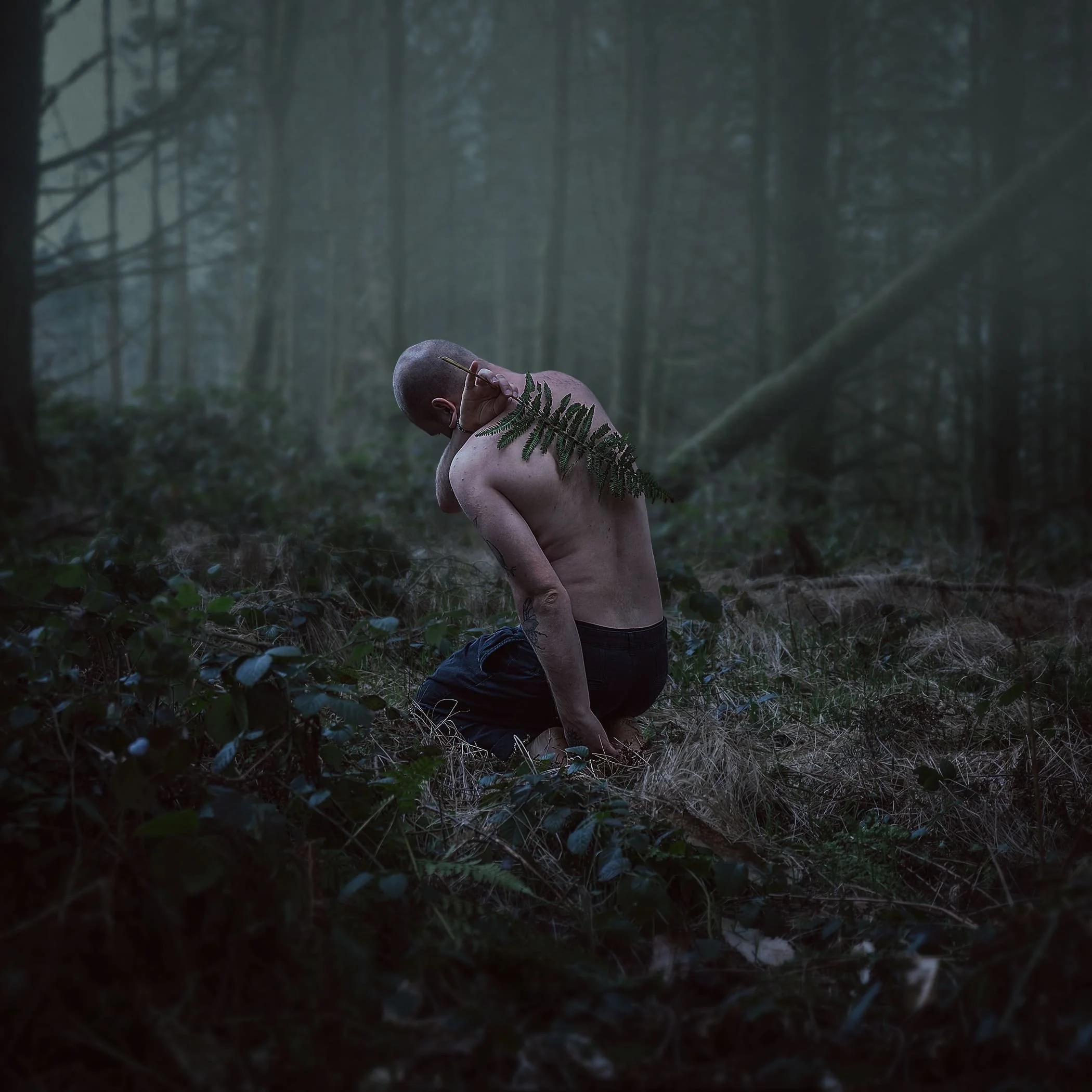 Ein nackter Mensch kniet in einem dunklen Wald mit einem Farnblatt auf der Schulter.