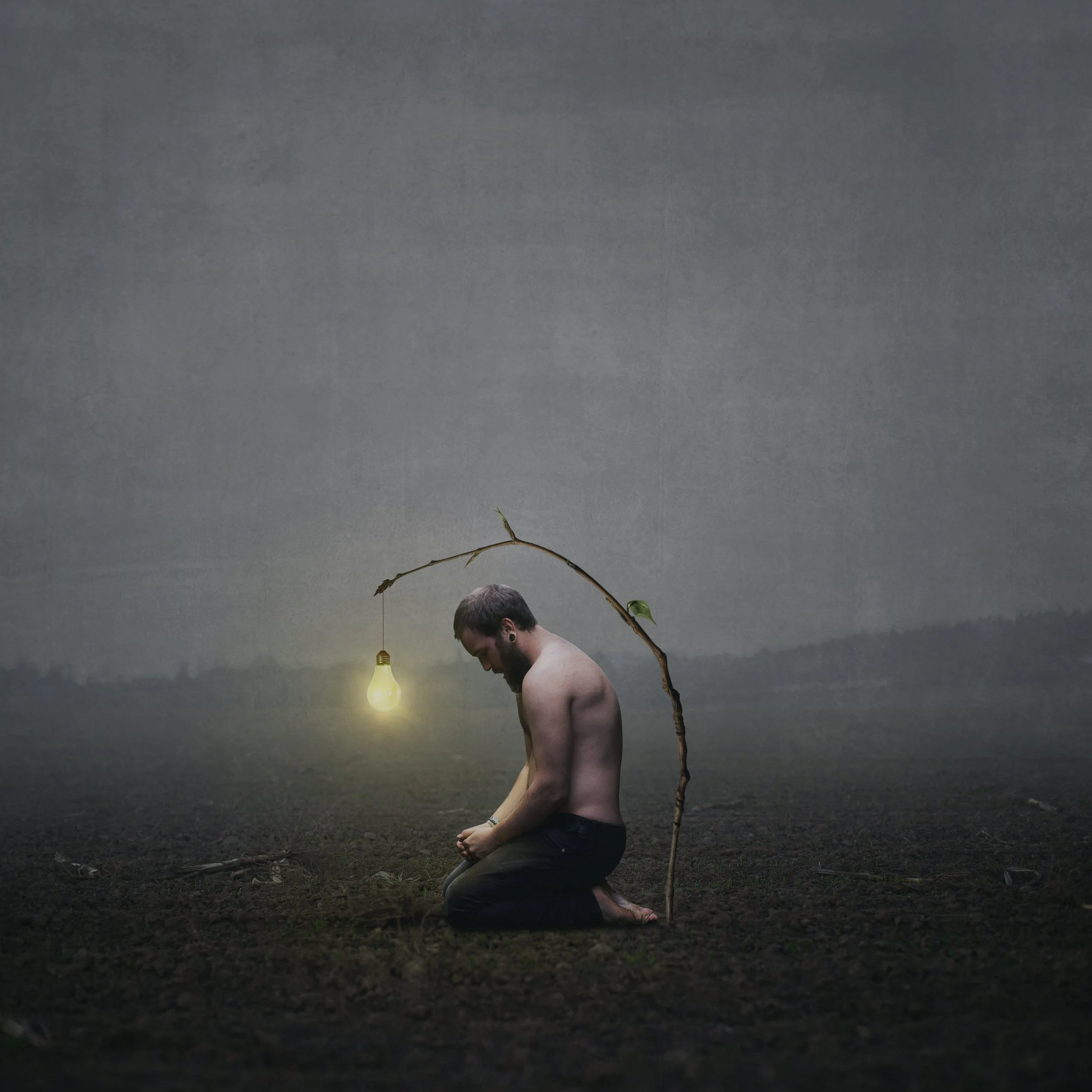Ein nackter Mann kniet mit gesenktem Kopf auf dunklem Boden, vor einem düsteren Himmel. Über seinem Kopf schwingt ein dünner Zweig mit einer leuchtenden Glühbirne, die an einem Faden hängt.