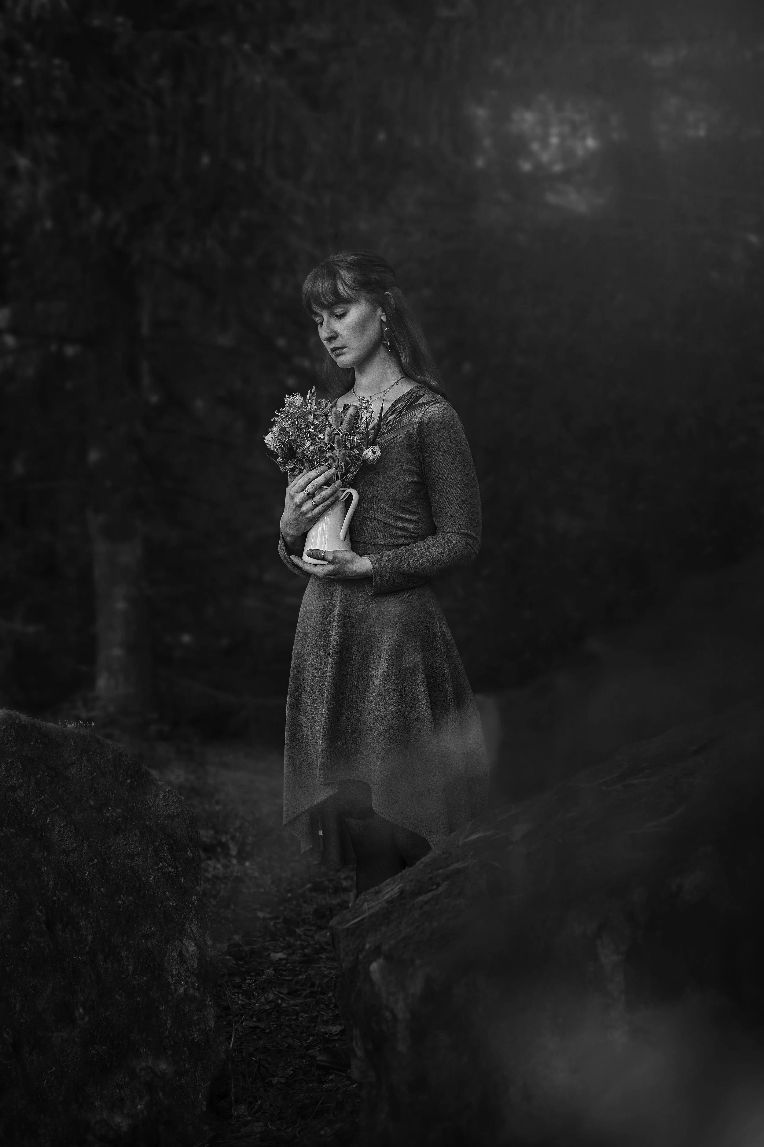 Schwarz-Weiß-Fotografie einer Frau, die in einem Wald steht und eine Vase mit Blumen hält, die Augen geschlossen hat.