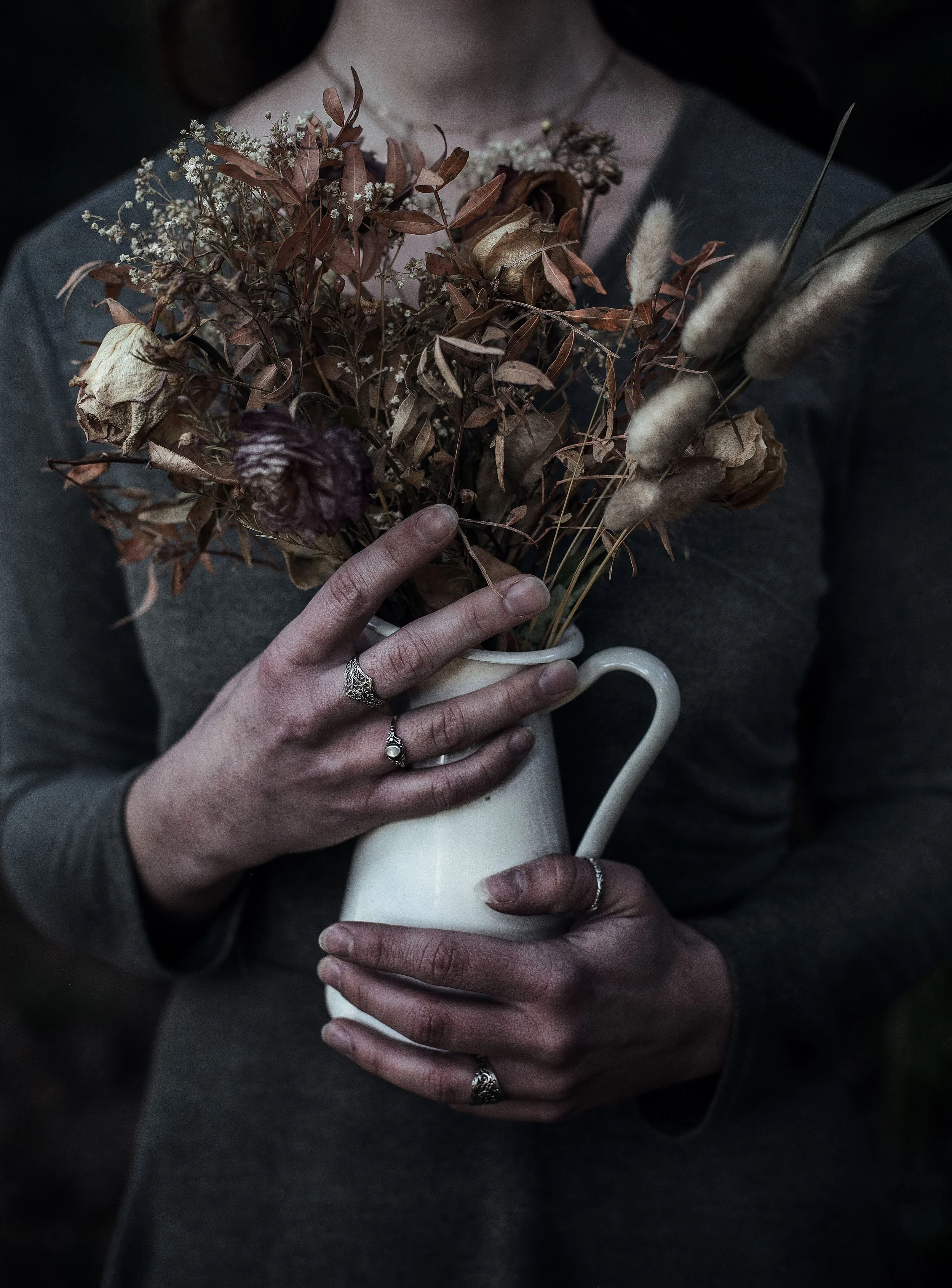 Person hält weiße Vase mit getrockneten Blumen, trägt Ringe an den Fingern, dunkle Kleidung, dunkler Hintergrund.