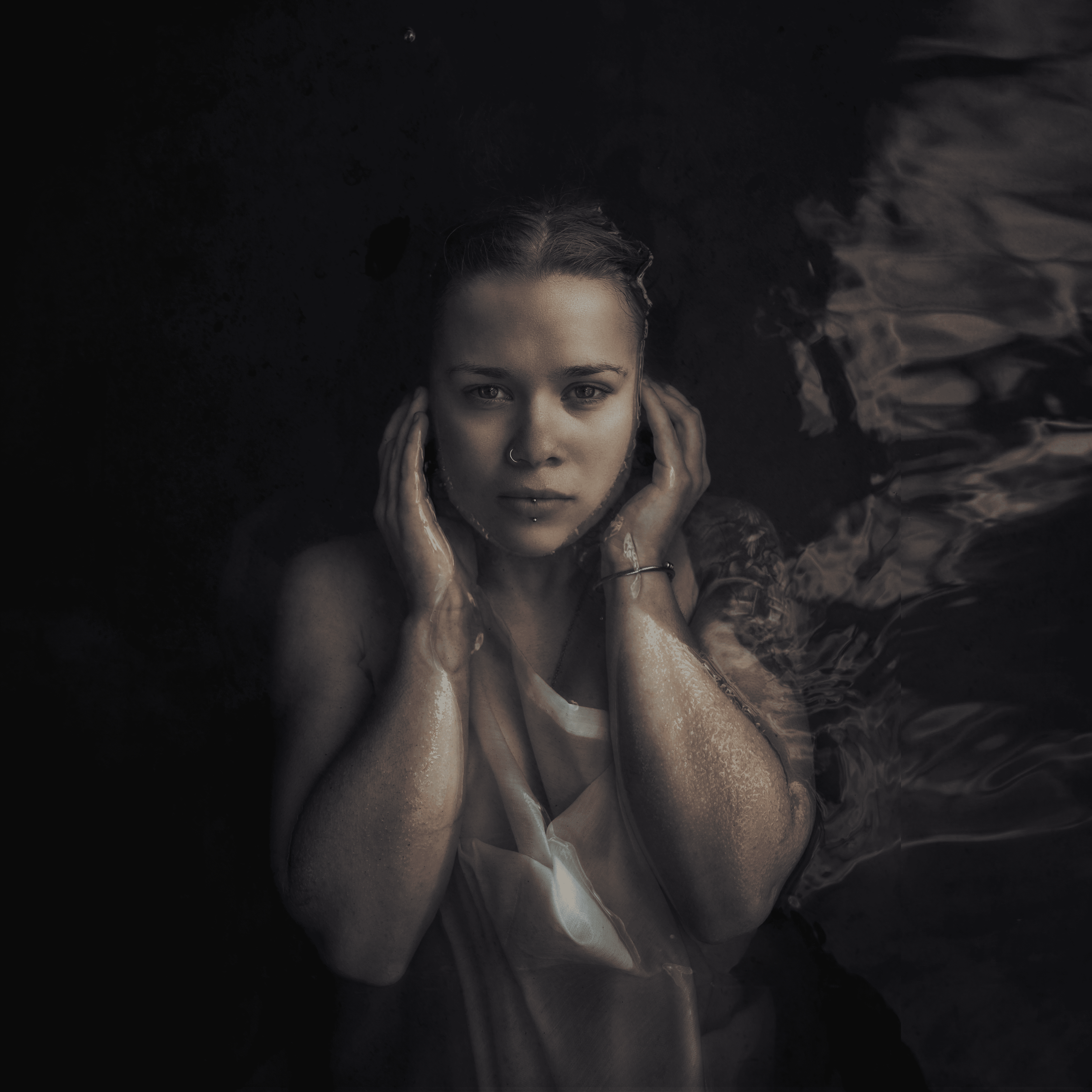 Junge Frau taucht im Wasser, hält ihre Hände an die Seite ihres Gesichts, schaut direkt in die Kamera, nackt nur mit einem nassen weißen Kleid. Dunkler Hintergrund mit Wasserreflexionen.
