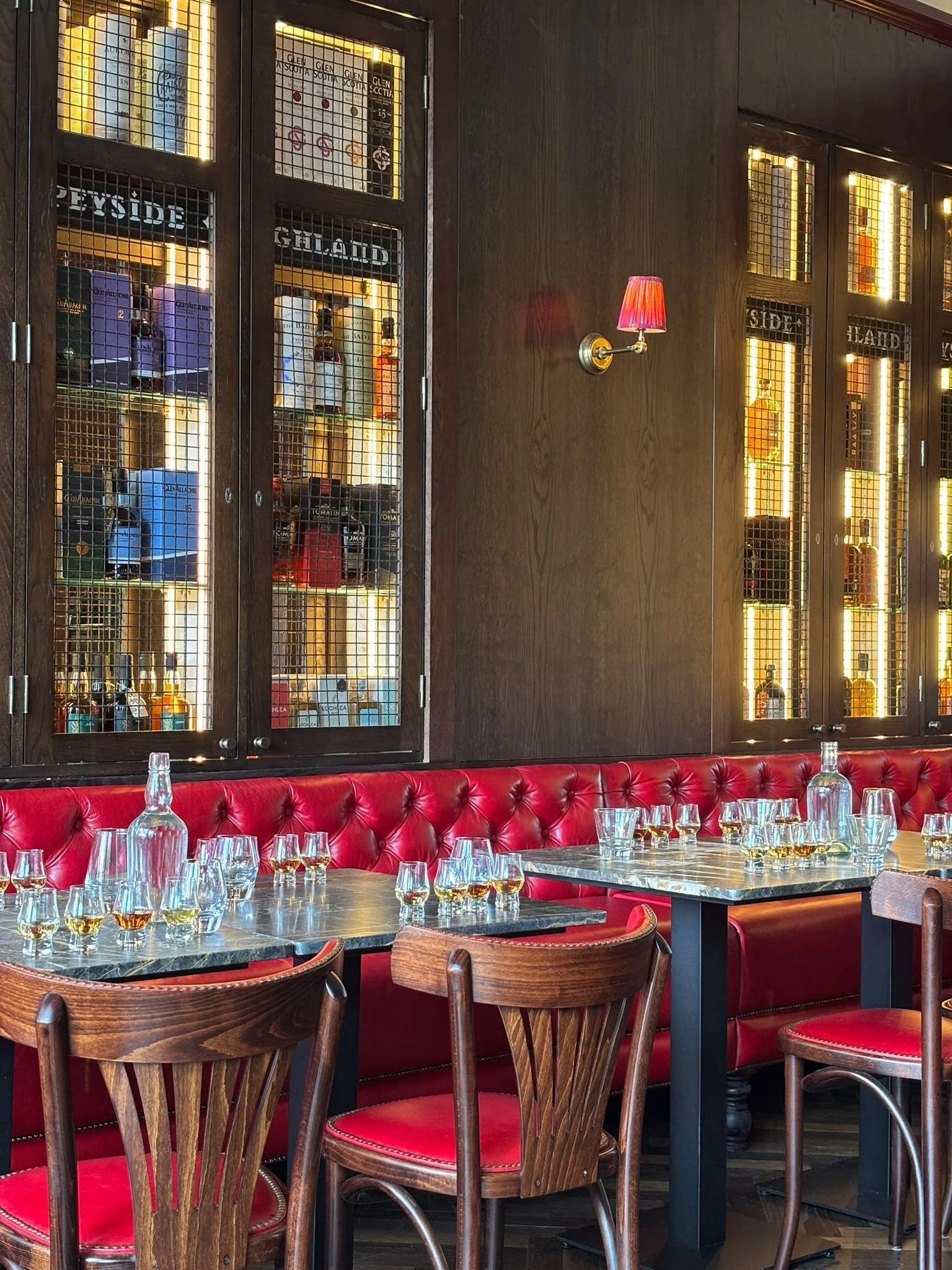 A cozy restaurant interior with dark wood paneling, glass-front liquor cabinets, red leather booth seating, and wooden chairs. Tables are set with glasses and bottles for drinks, illuminated by warm wall sconces.