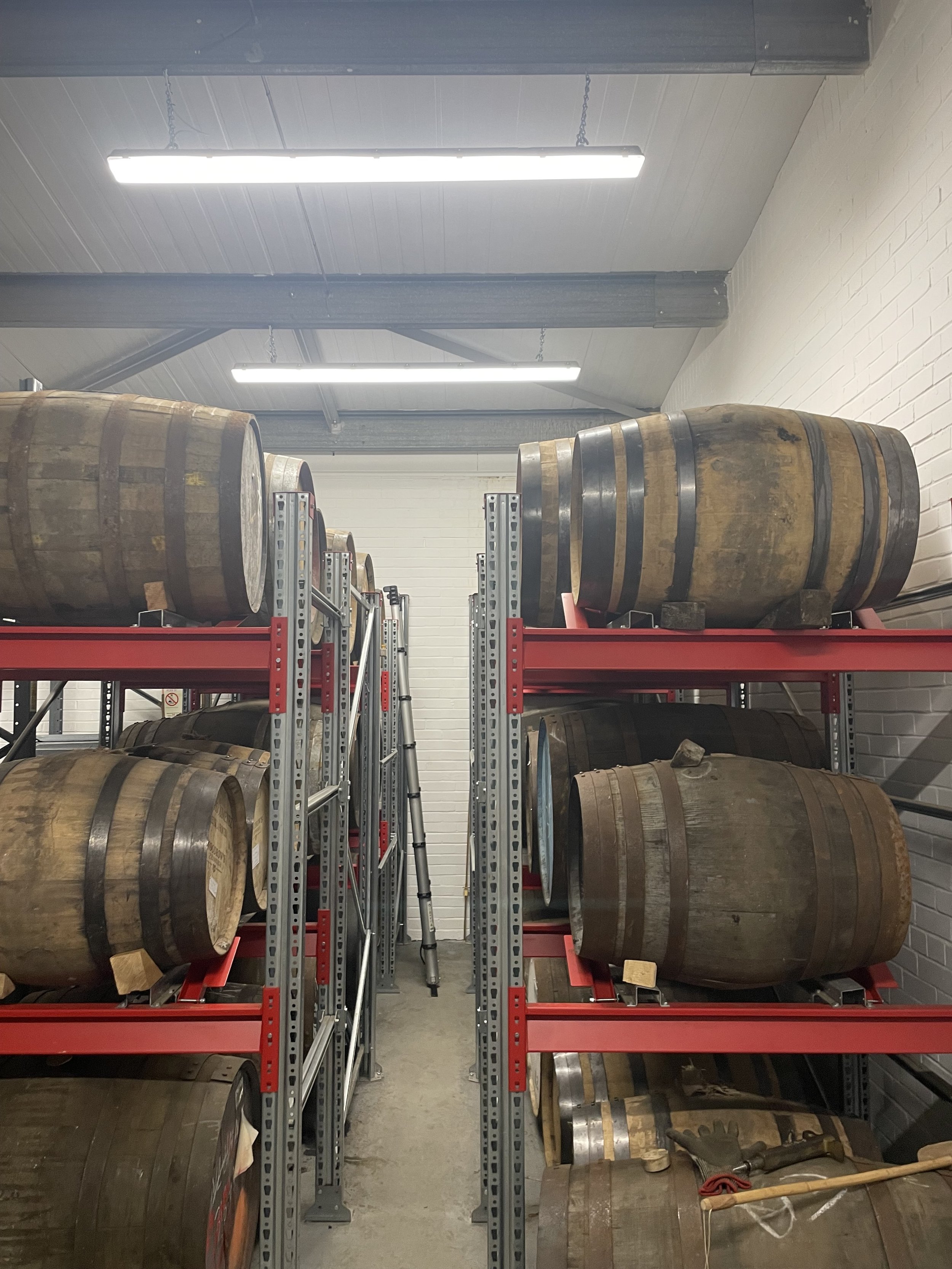 Storage room with metal shelves holding whiskey barrels, some on the floor and others on the shelves, with various tools and equipment nearby.