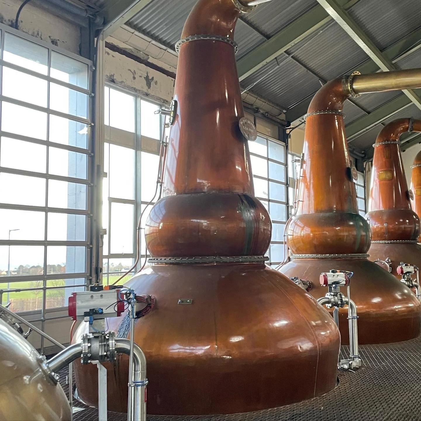 Large copper stills used in whiskey production inside a distillery, with tall windows allowing natural light